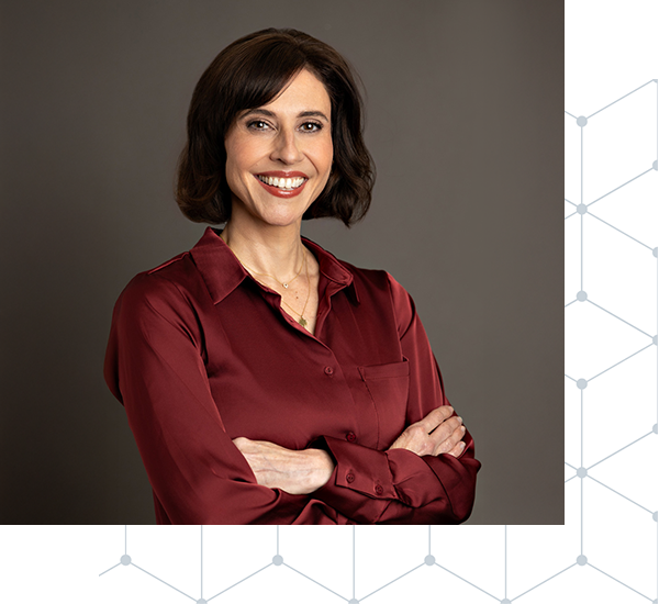 Portrait of a woman with shoulder-length dark hair, wearing a maroon silk blouse, smiling with arms crossed against a gray background with a partial geometric pattern on the right side.