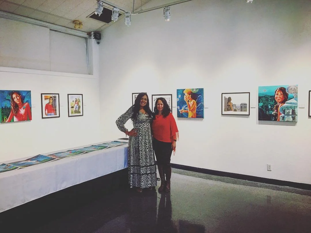 Two women standing in an art gallery, smiling, with framed and unframed artwork on the white walls and a table with laying artwork.
