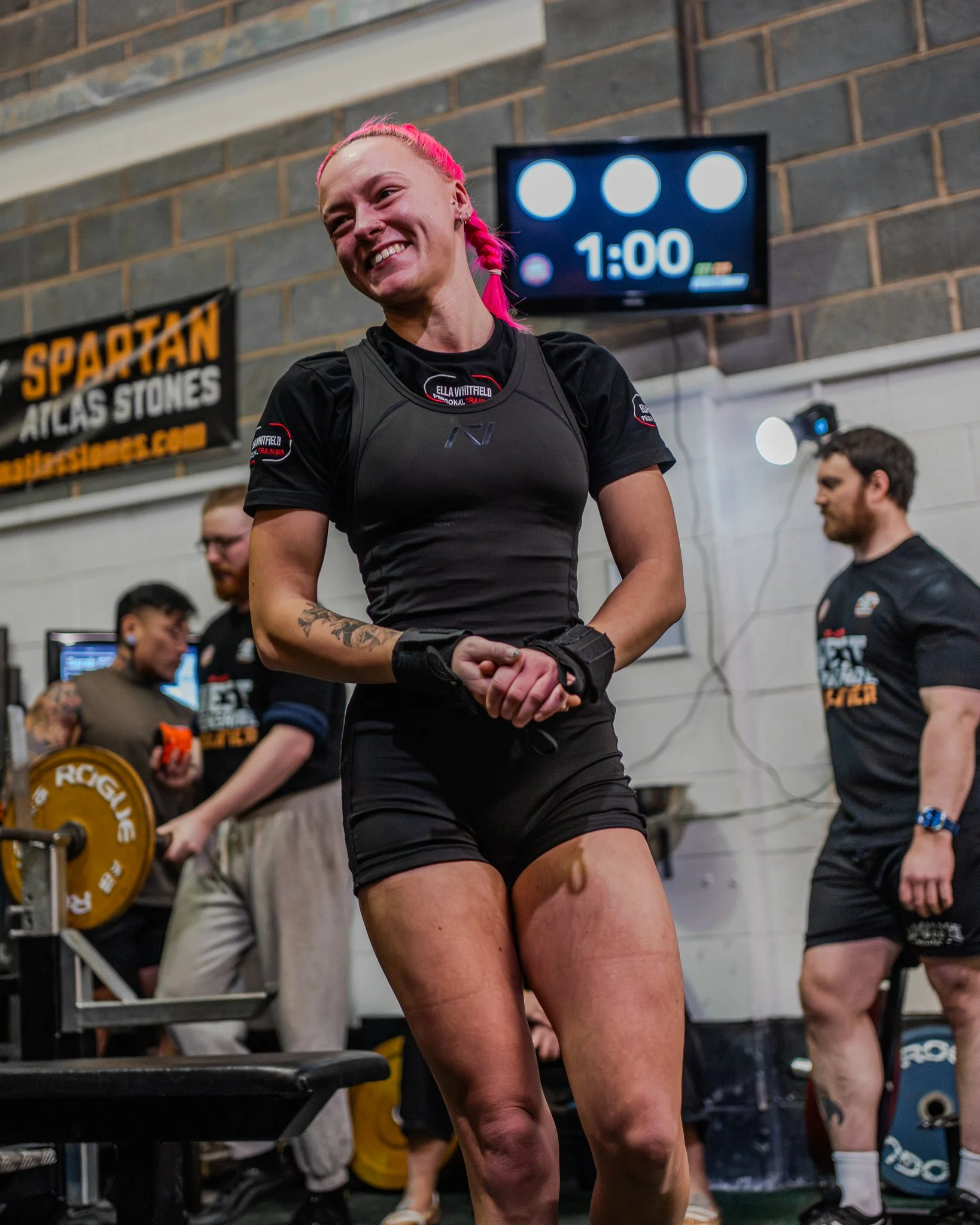 A woman with pink hair, smiling, in a black athletic outfit, in a gym with a digital timer showing 1:00 in the background. Other people are present, including a man with a beard and black shirt and another person lifting weights.