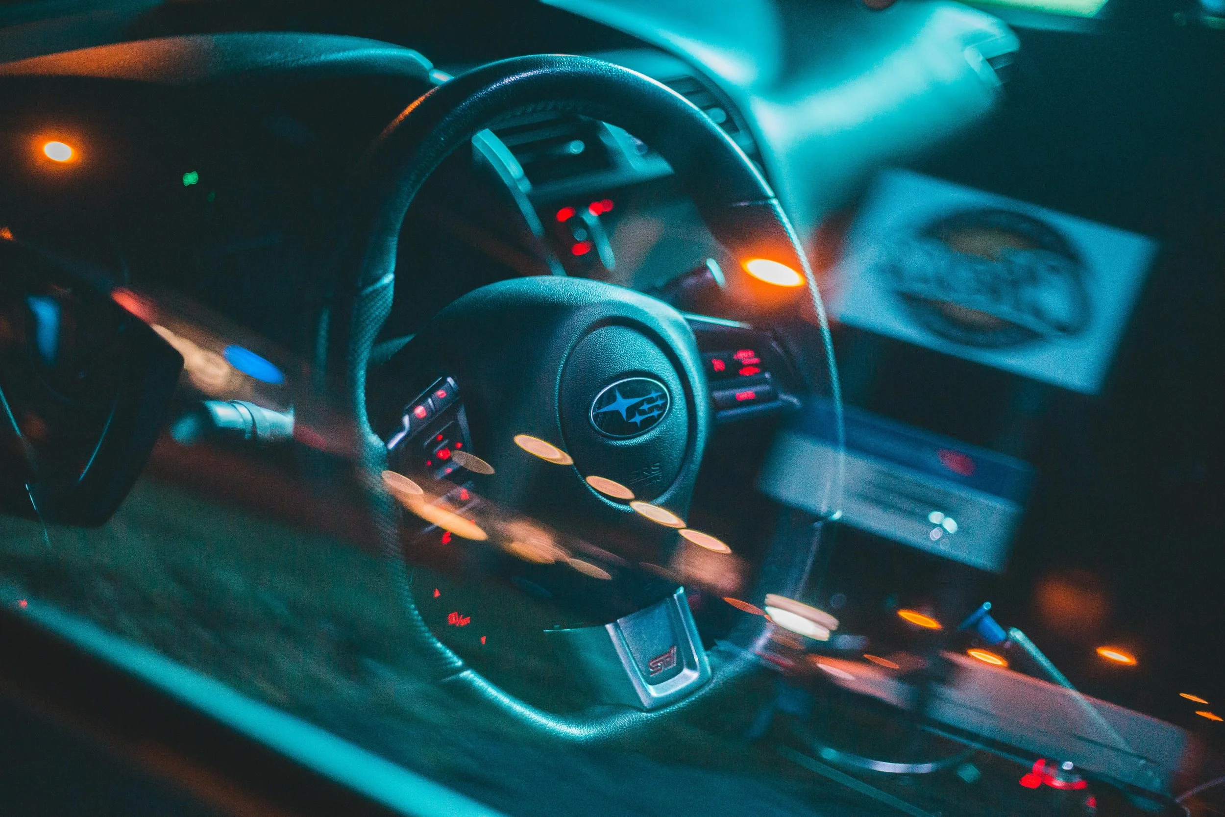 Interior of a car showing the steering wheel with Subaru logo, dashboard with illuminated controls, and a display screen, all illuminated by orange and blue lights.