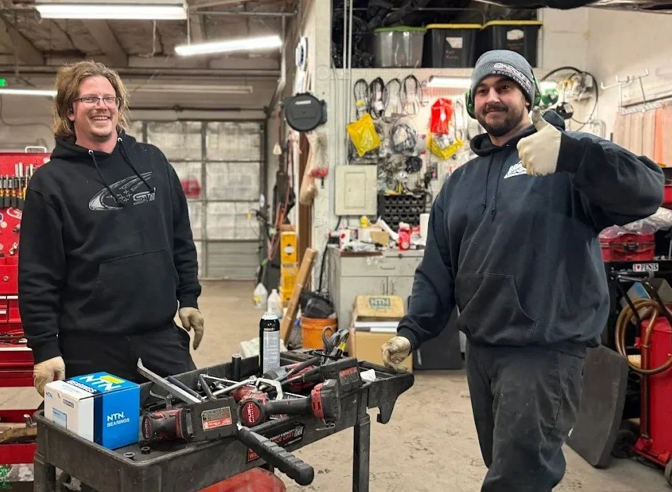 Two men in black hoodies and gloves standing in a workshop. One man is smiling, the other giving a thumbs-up. They are near a workbench with tools and supplies, with various equipment and storage on the walls behind them.