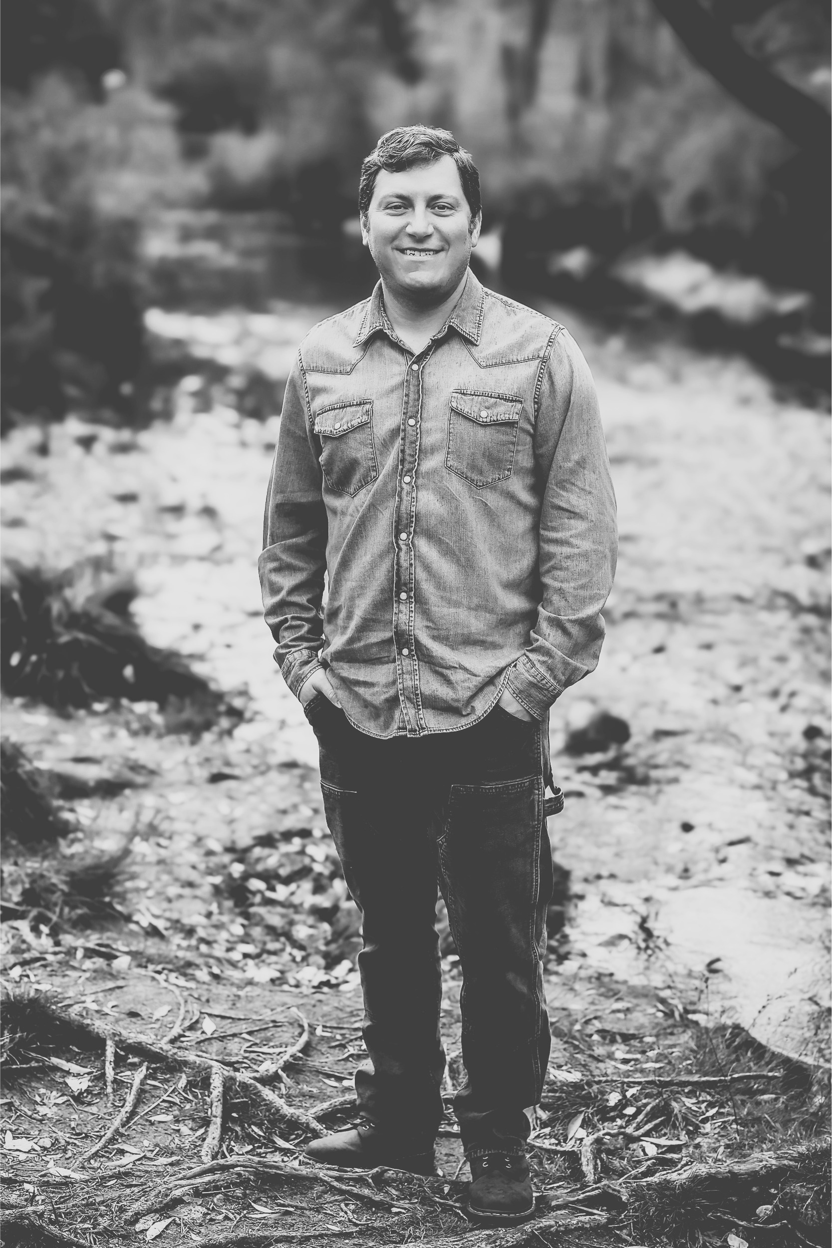Black and white photograph of a young man standing outdoors on a trail covered with leaves and twigs. The background features blurred trees and natural scenery.