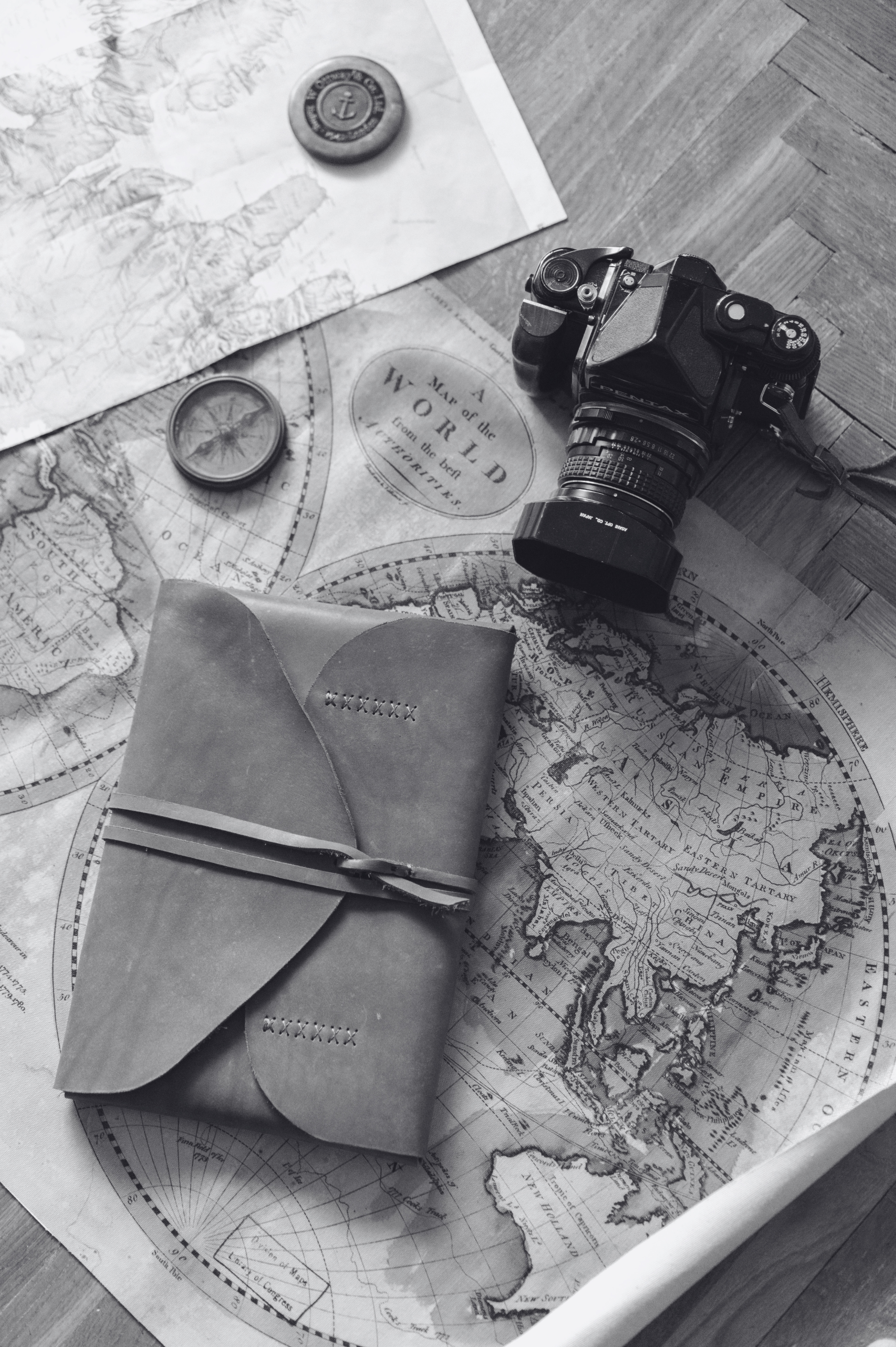 Black and white photo of a vintage camera, two compasses, a map, and a journal with stitched X markings, all placed on a wooden surface, suggesting an exploration or travel theme.