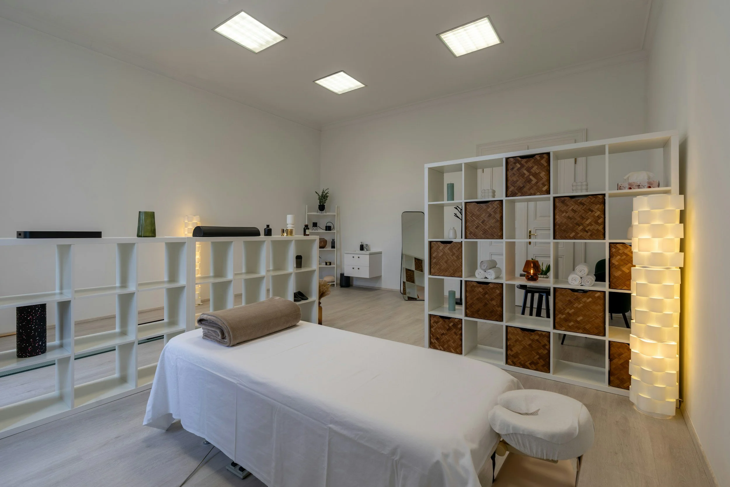 A massage therapy room with a massage table covered in white sheets, a folded towel at the head, and a massage stool nearby. The room features modern shelving with decorative items, a standing mirror, and a soft warm lighting lamp, creating a calm atmosphere.