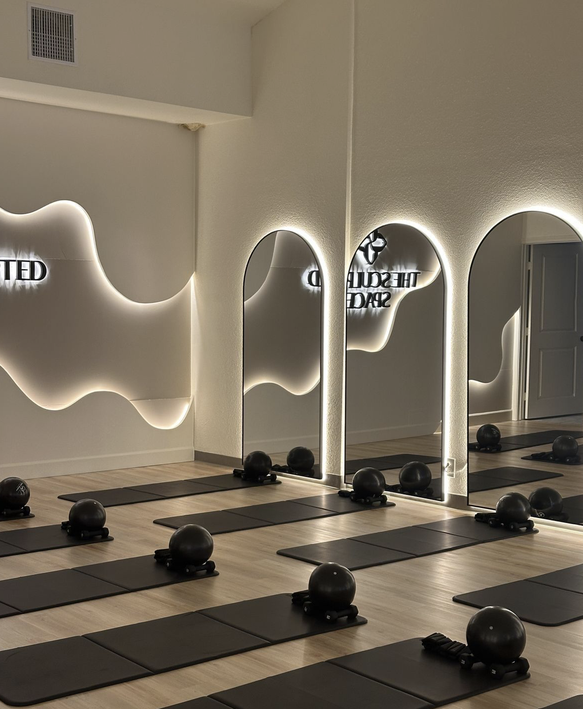 Fitness studio with black mats, medicine balls on small stands, and mirrors with backlit edges on the wall.