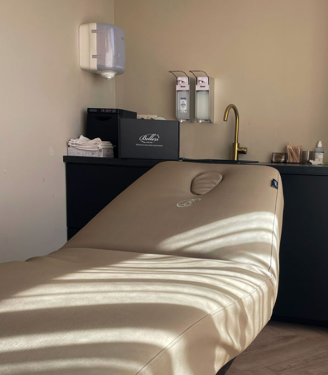 Medical examination room with a beige massage or exam table, a sink with a gold faucet, and various medical supplies and equipment on a black countertop against a beige wall.