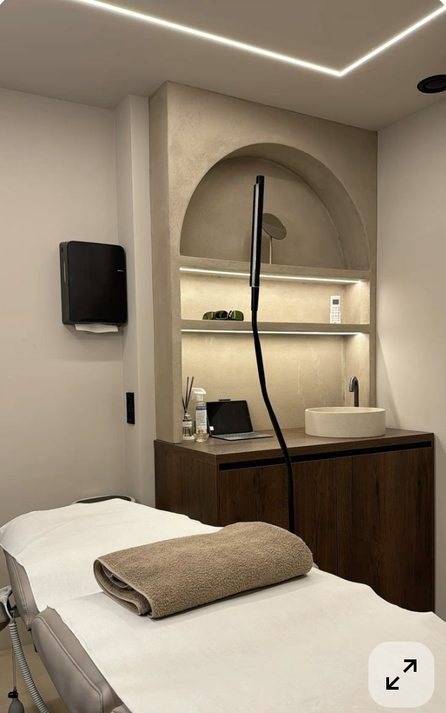 A massage room with a massage table covered by a towel and a rolled-up towel on top. There is a dark wooden cabinet with a sink, a small mirror, skincare products, and a tablet. Behind the cabinet, an arched wall niche with shelves illuminated by LED lighting holds a phone and some objects. A wall-mounted speaker is on the wall, and the ceiling has modern LED lighting.