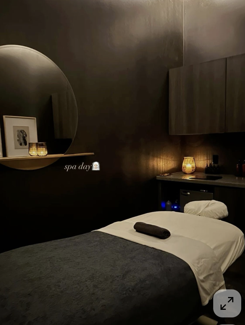 A dimly lit spa room with a massage table covered in dark gray blanket, a rolled towel on top, and a white sheet hanging over the side. There is a small counter with a lit decorative lamp, bottles, and a mini fridge beneath. A mirror and framed artwork are on the dark wall, and a circular shelf holds candles and a framed photo. The overlay text says 'spa day' with a spa emoji.