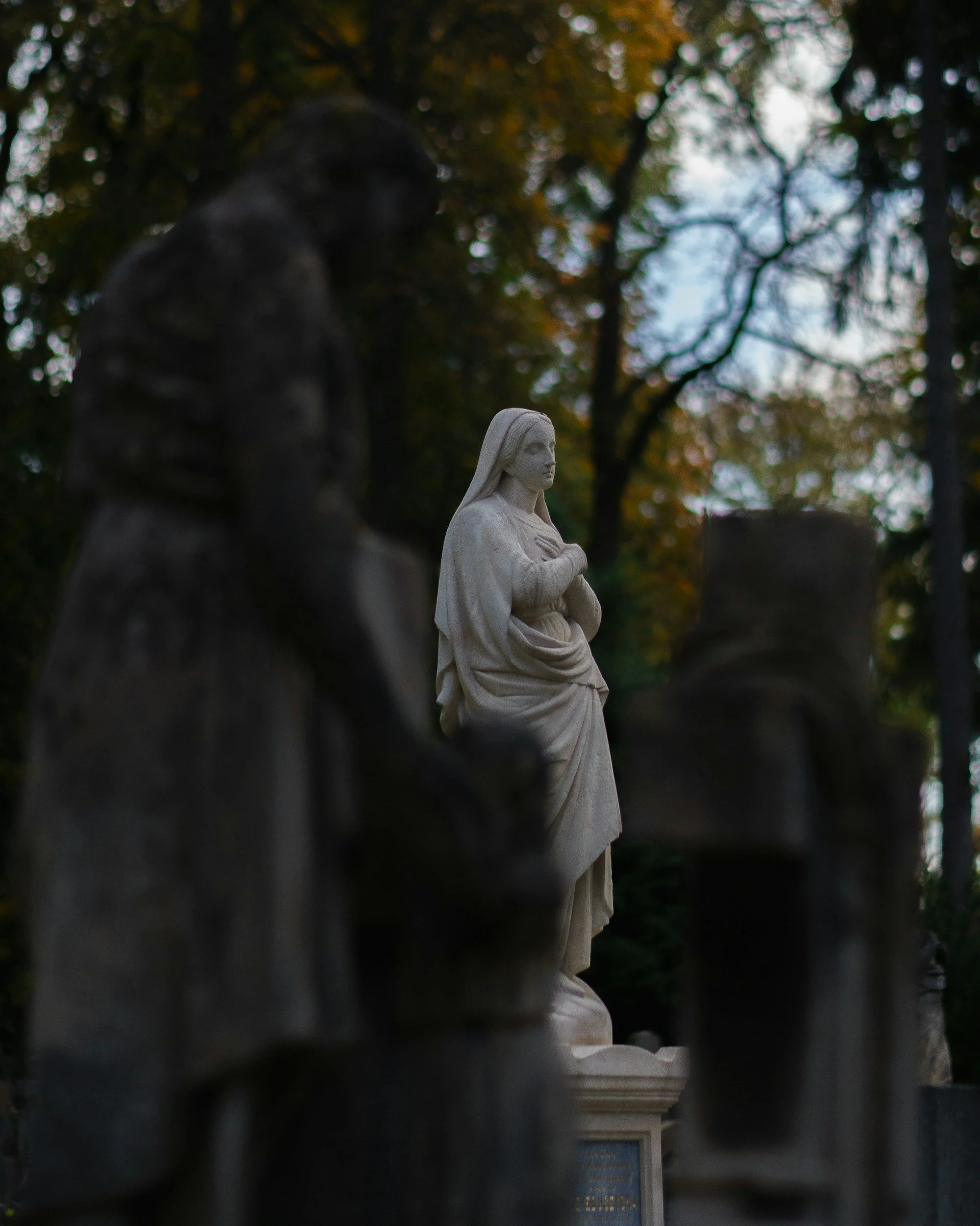 A white marble statue of a woman with long hair, standing with her hands crossed over her chest, surrounded by smaller dark statues, with trees and autumn leaves in the background.