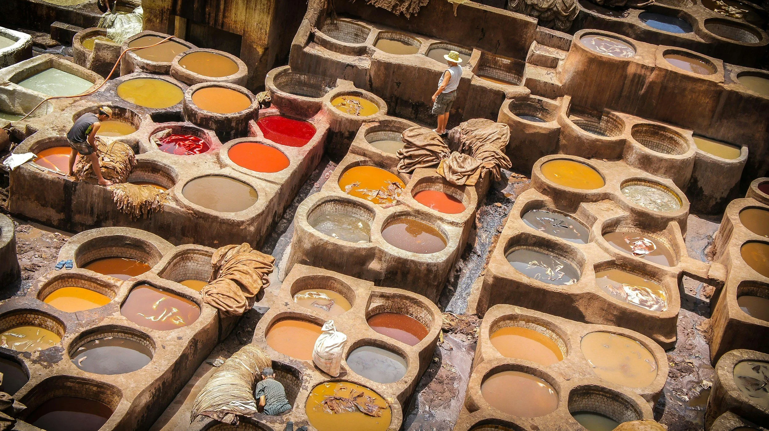 Workers standing and sitting among large stone vats filled with colorful liquids, likely for traditional dyeing or processing, in an outdoor workshop setting.