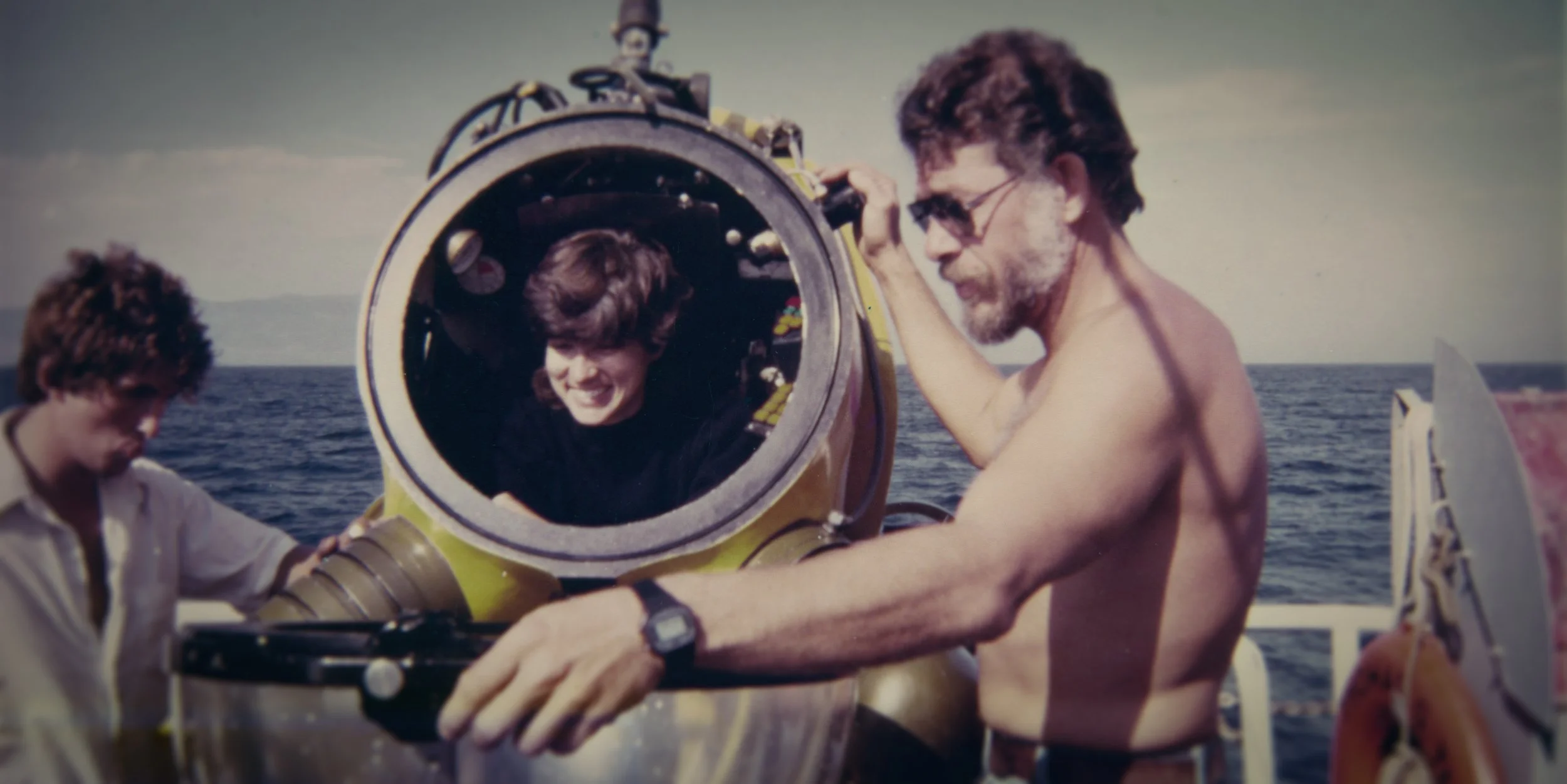 A man with sunglasses and a beard is shirtless and working with a large underwater scooter. A woman is sitting inside the scooter's cockpit, smiling. A younger person is standing next to the man. The scene takes place on a boat during daytime with open water and a distant shoreline in the background.