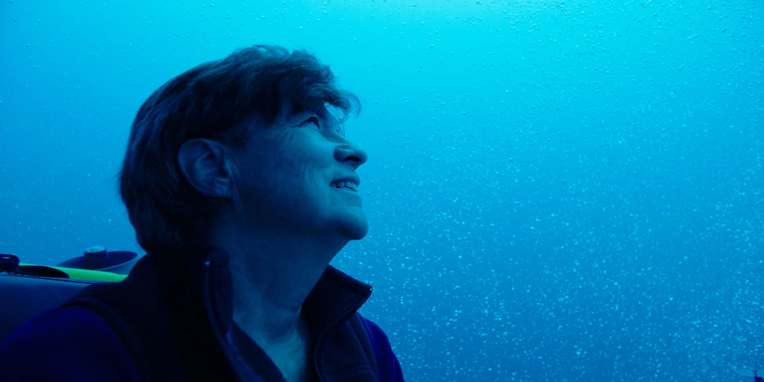 A woman with short brown hair, smiling, looking up through rain-covered window.