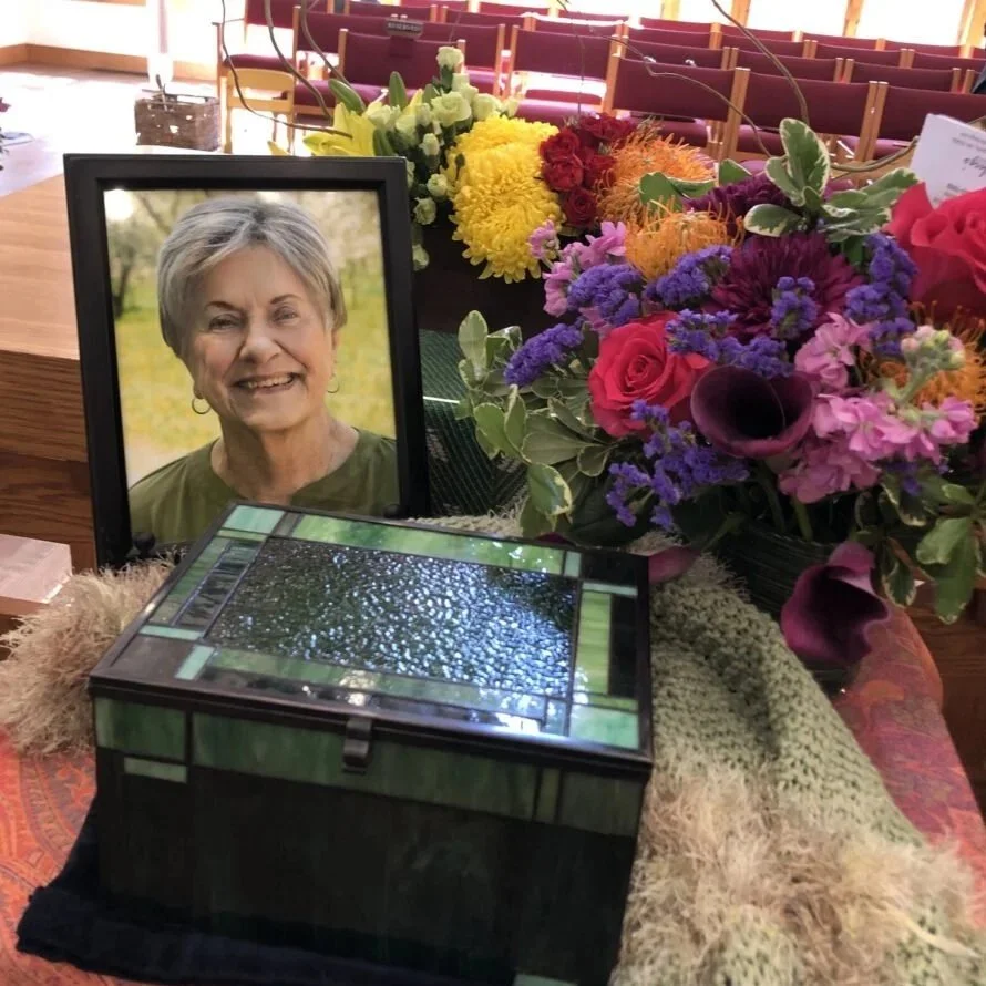 A memorial setup with a framed photograph of a woman, surrounded by flowers and a decorative box, on a table with empty red seats in the background.