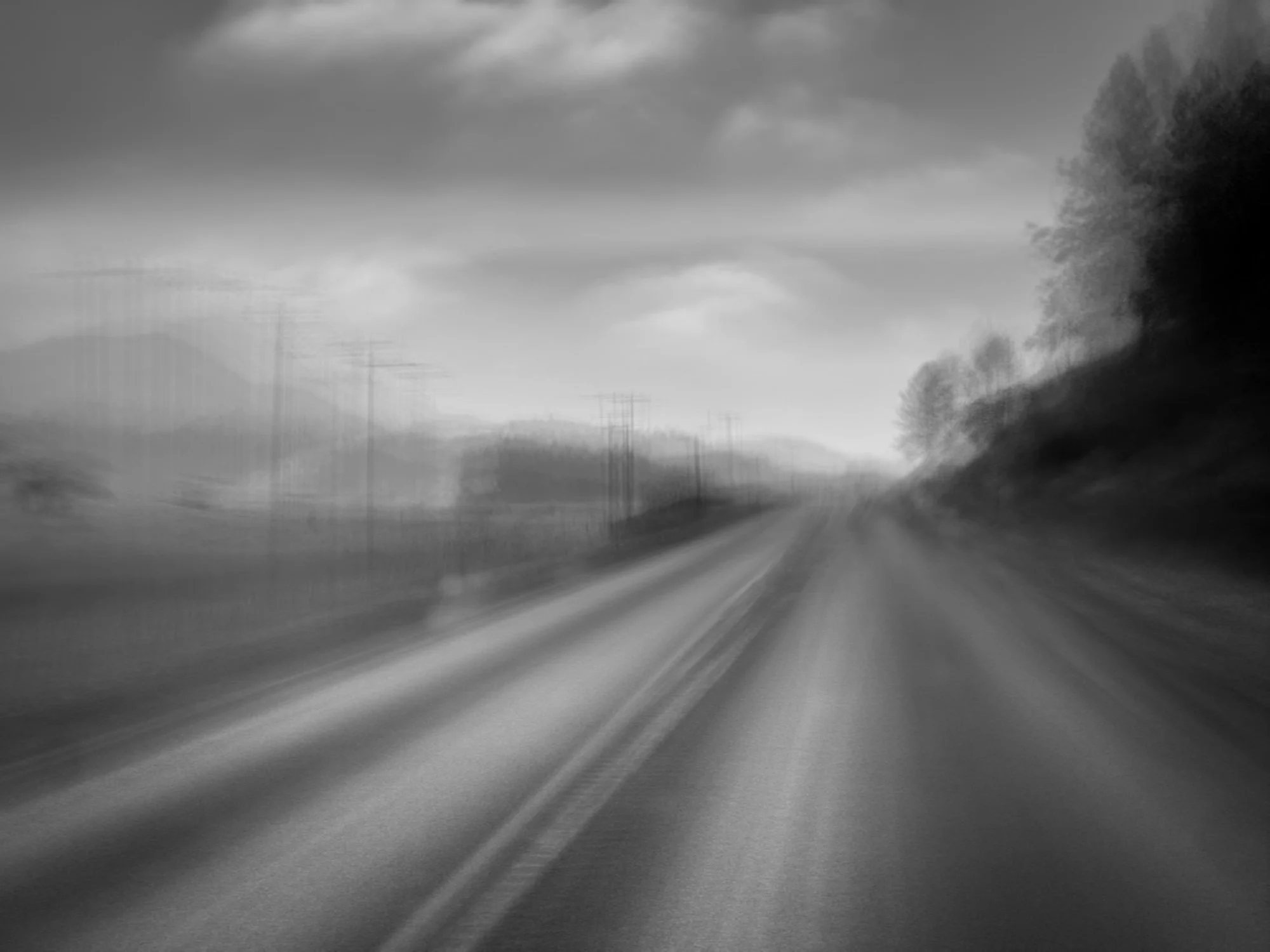 Black and white photo of a straight road with blurred motion effect, trees on the right, power lines on the left, cloudy sky overhead.