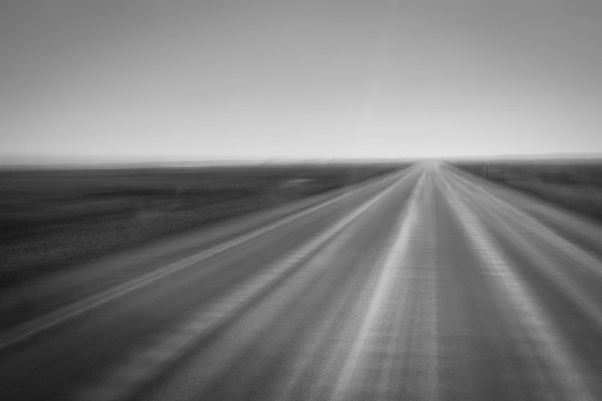 A blurred black-and-white photo of a straight road stretching into the horizon in a flat, open landscape.