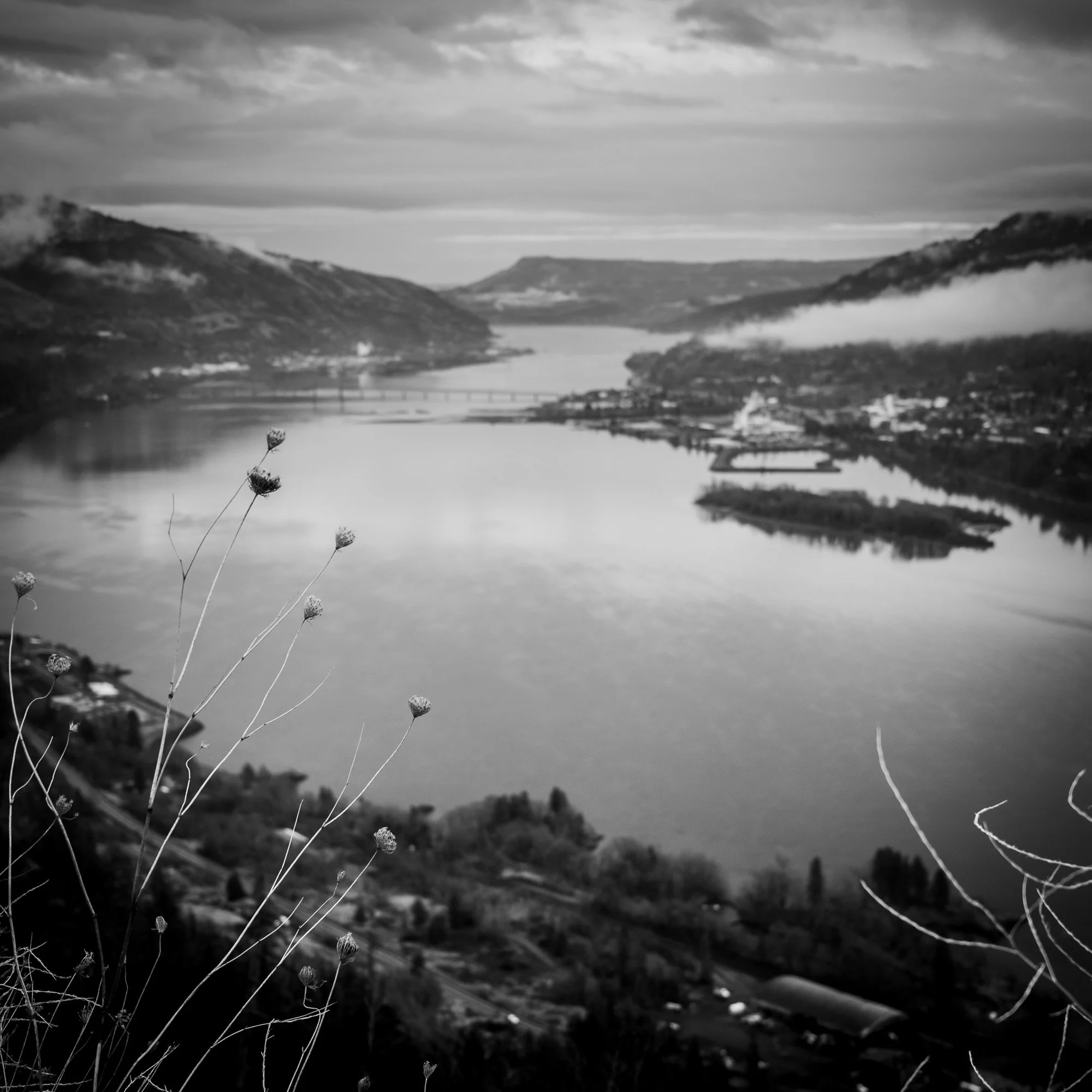 Black and white photo of a wide river flowing through a valley with hills on either side, and a cloudy sky overhead.