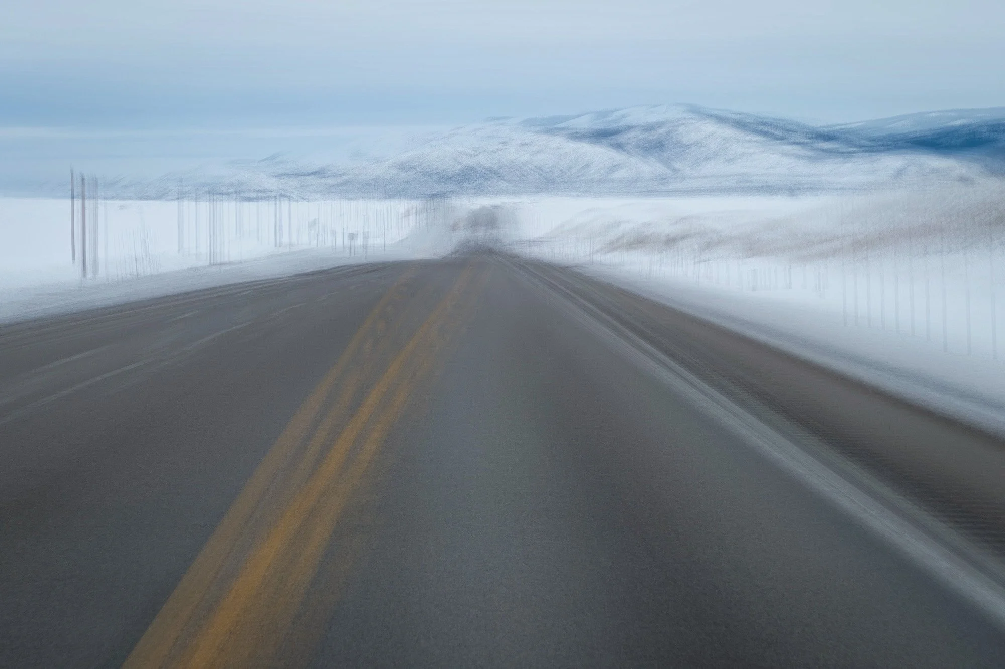 Blurred image of a two-lane road with yellow center lines, surrounded by snow-covered fields and mountains in the background.