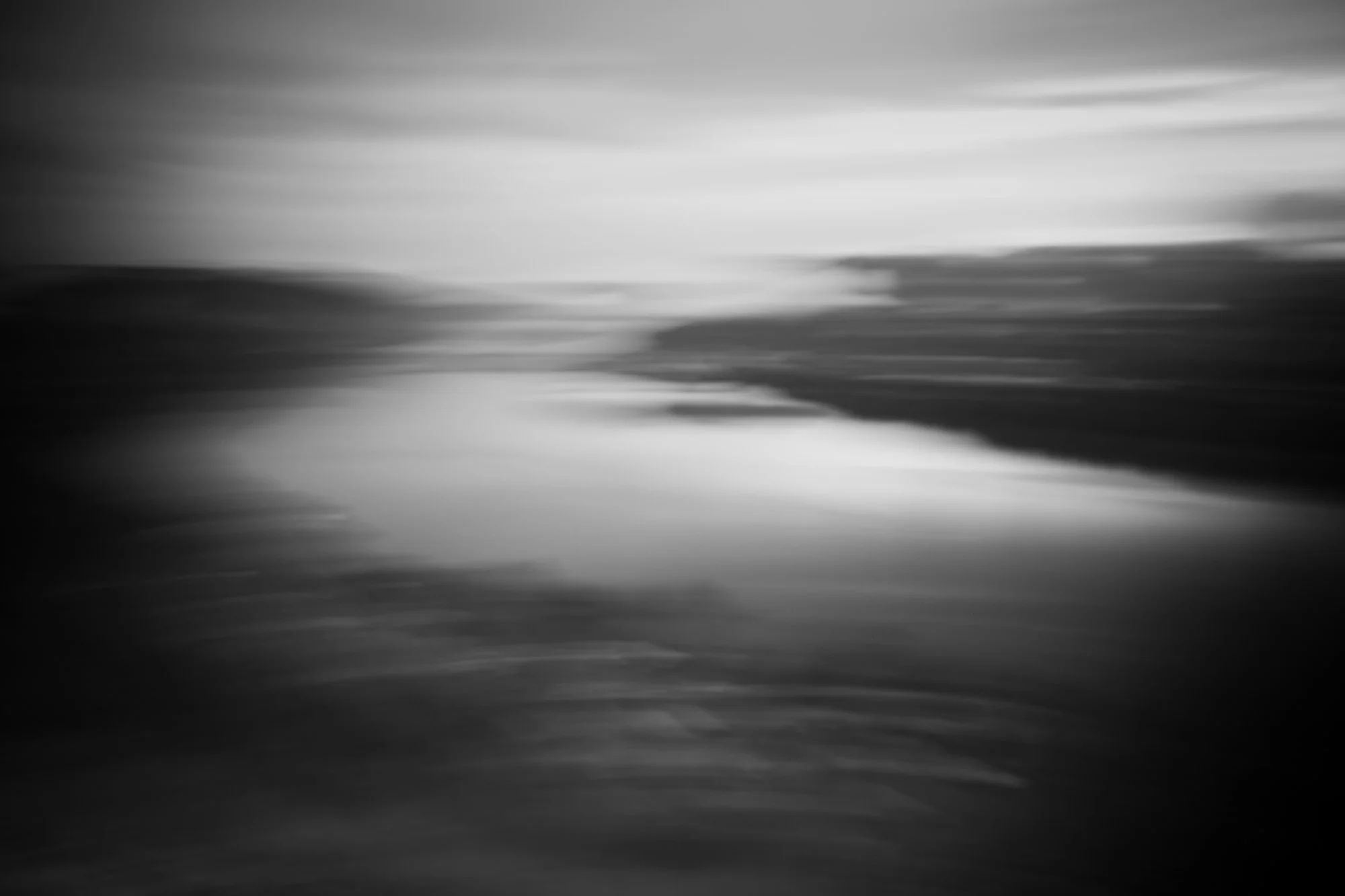 Blurry black and white photo of a body of water with a distant shoreline and cloudy sky.