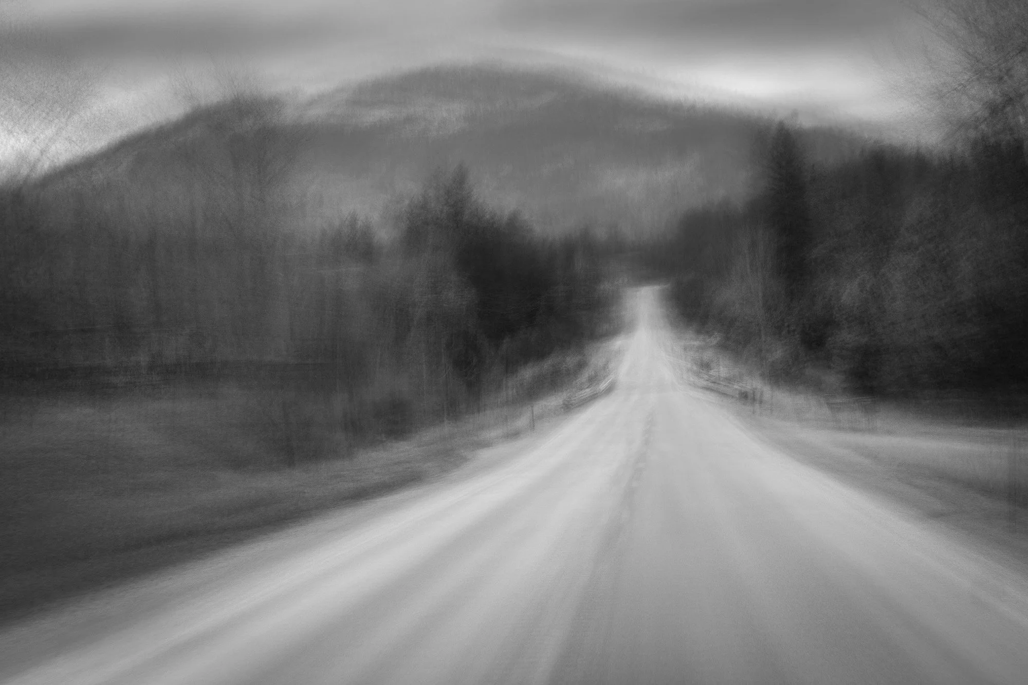 Blurred black and white photo of a long, straight road with trees on both sides and hills in the background.