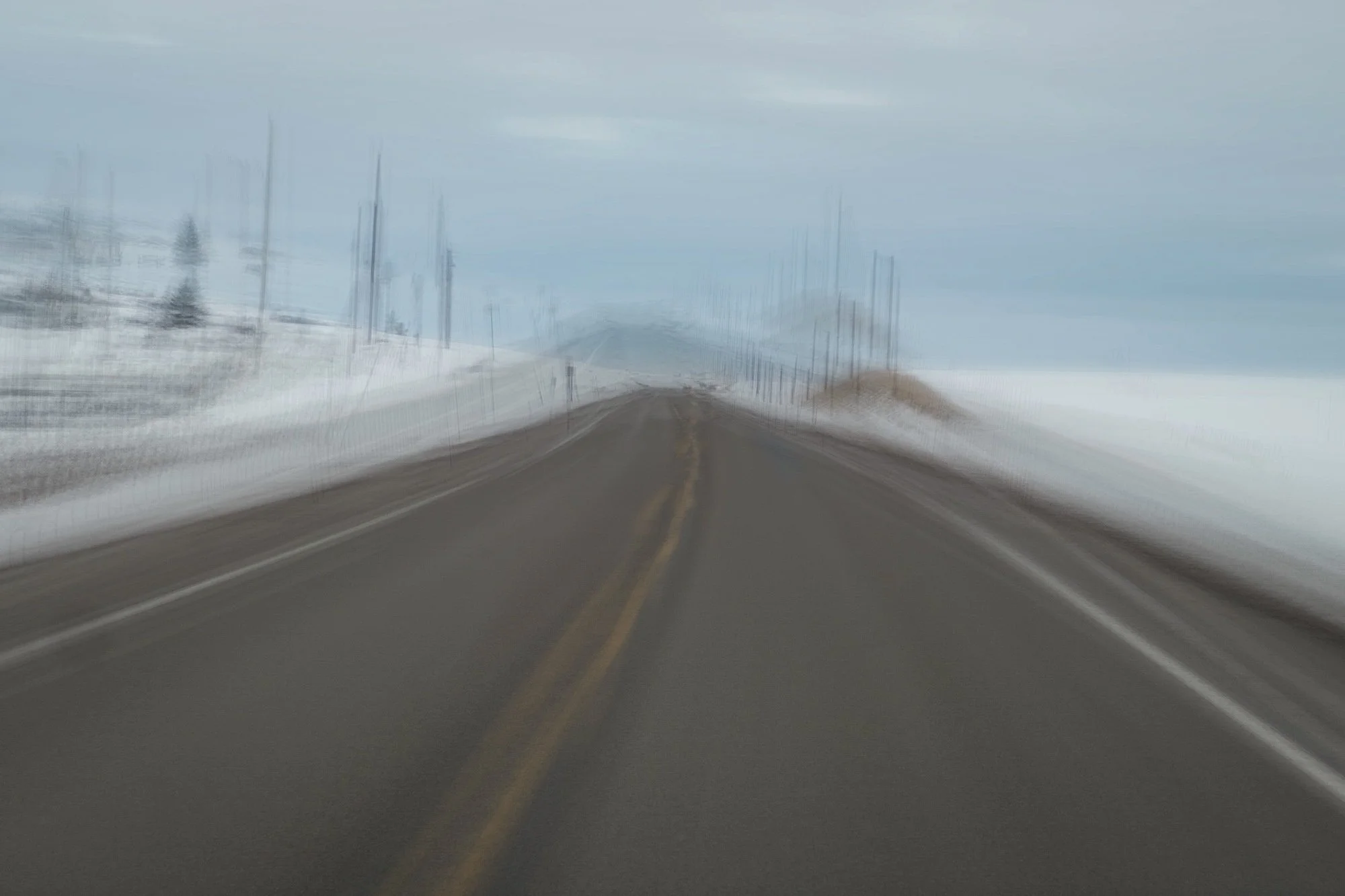 Blurred photo of a rural road with snow on sides, power lines and trees in the distance, under an overcast sky.