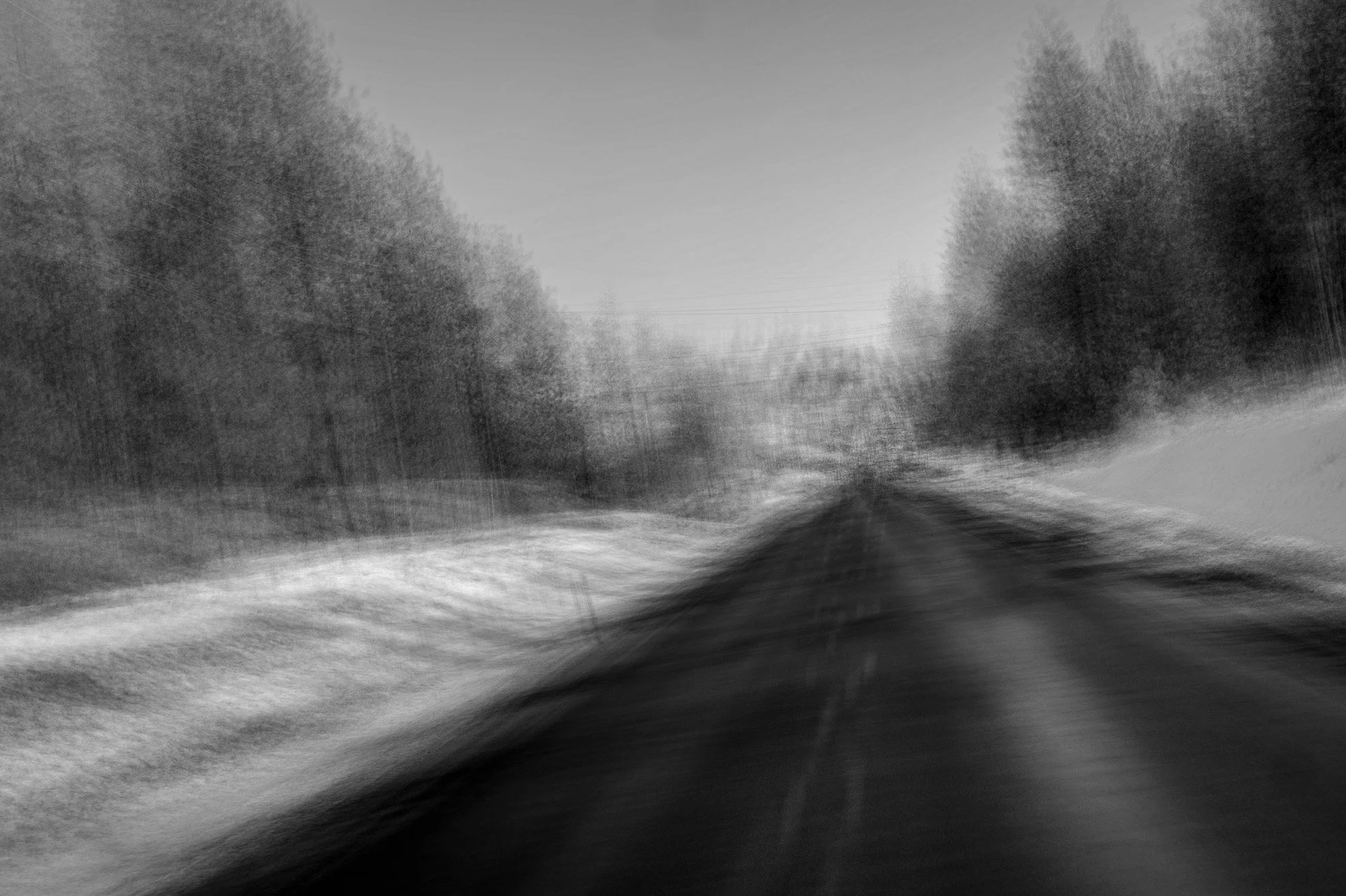 Blurry black and white photo of a straight road surrounded by trees on both sides, perceived movement or motion blur.