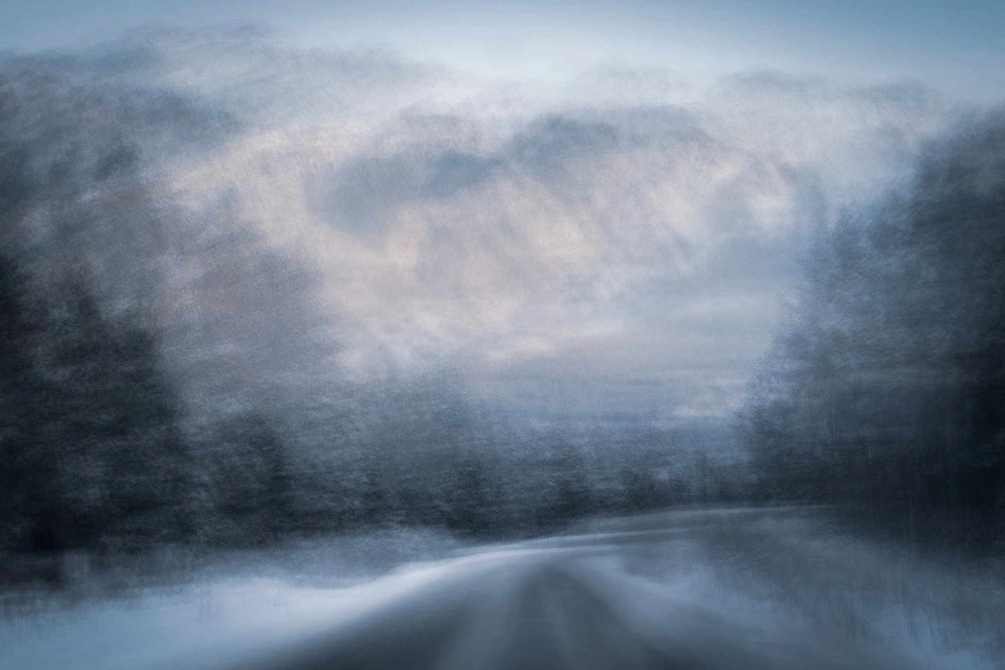 Blurry image of a snow-covered landscape with trees in the distance and a road or path in the foreground, under a cloudy sky.