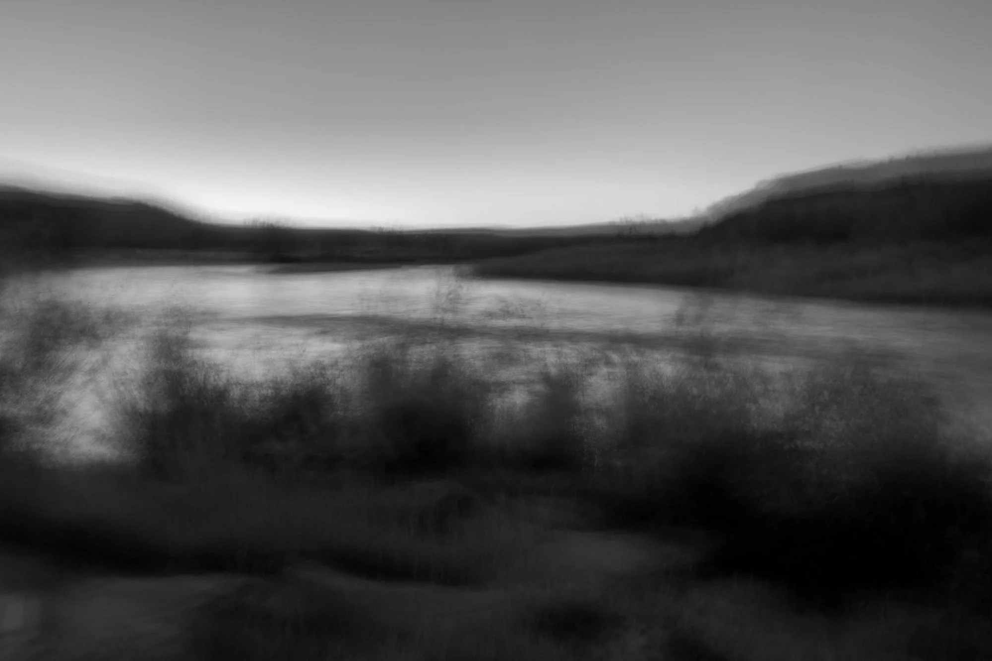 Black and white photo of a landscape with a river, hills, and a sky at sunset or sunrise, appearing blurry and abstract.
