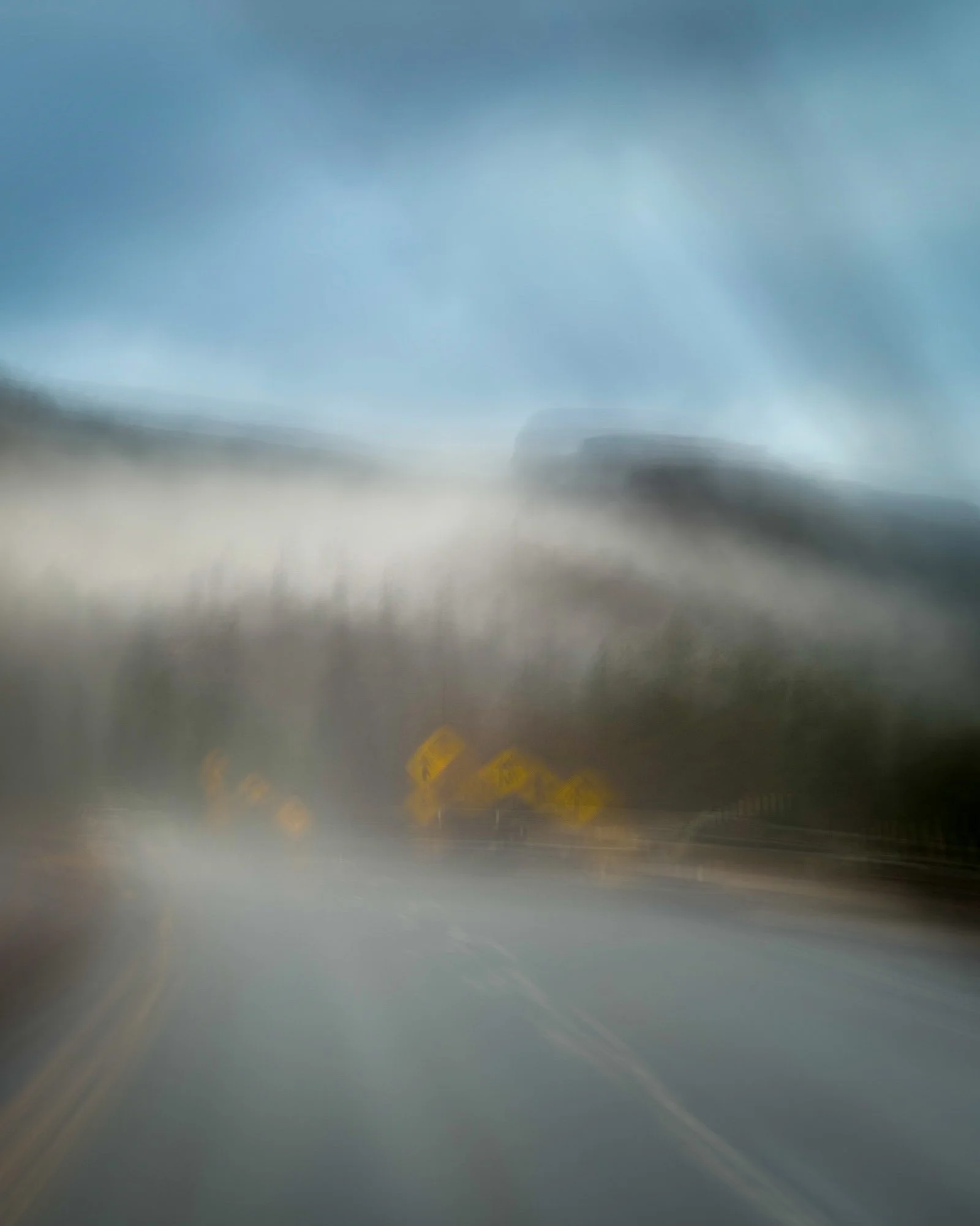 Blurry image of a highway with yellow road signs and a faint outline of a vehicle in the distance, under a cloudy sky.