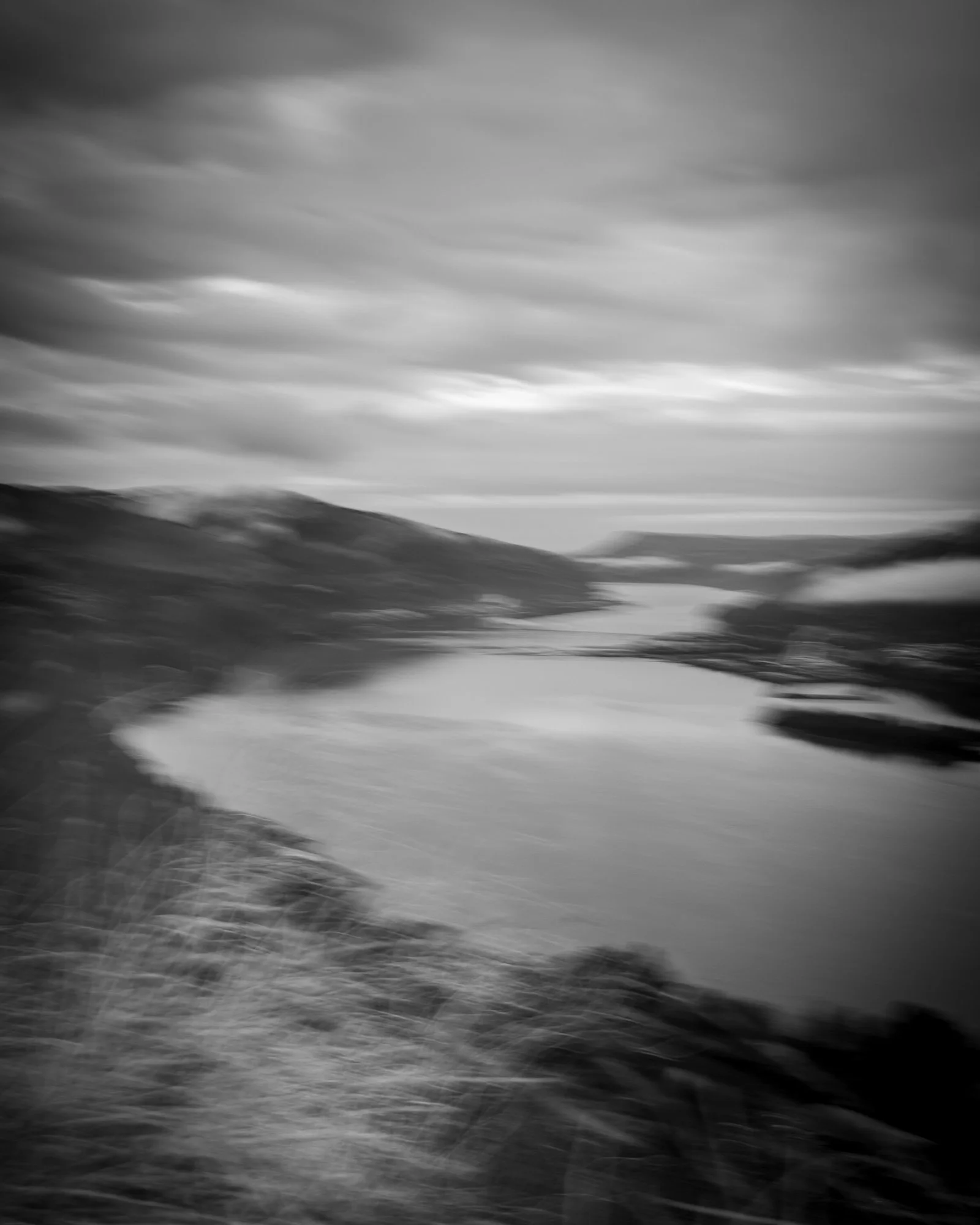 A black and white photograph of a river meandering through a landscape with mountains in the background and a cloudy sky overhead.