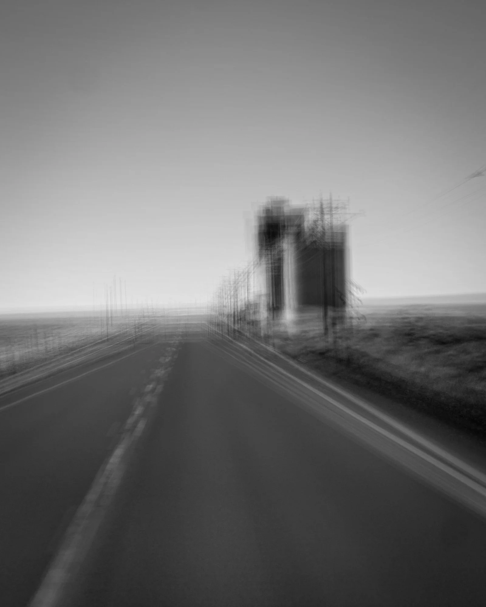 A black and white photograph of a rural road with motion blur, flanked by fields and a few buildings in the distance.