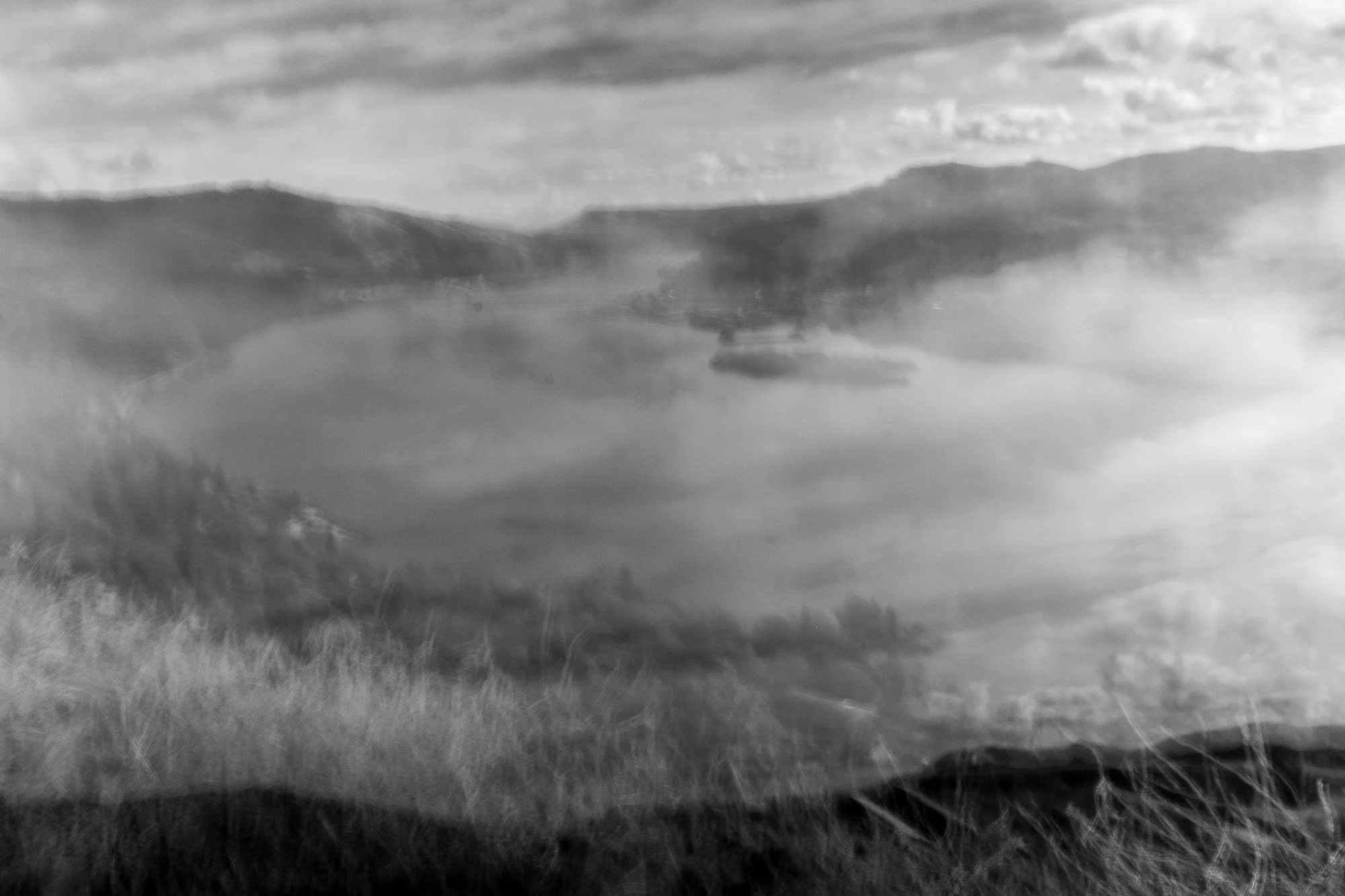 Black and white photo of a lakeside scene with mountains in the background, reflecting in the water, and a cloudy sky overhead.