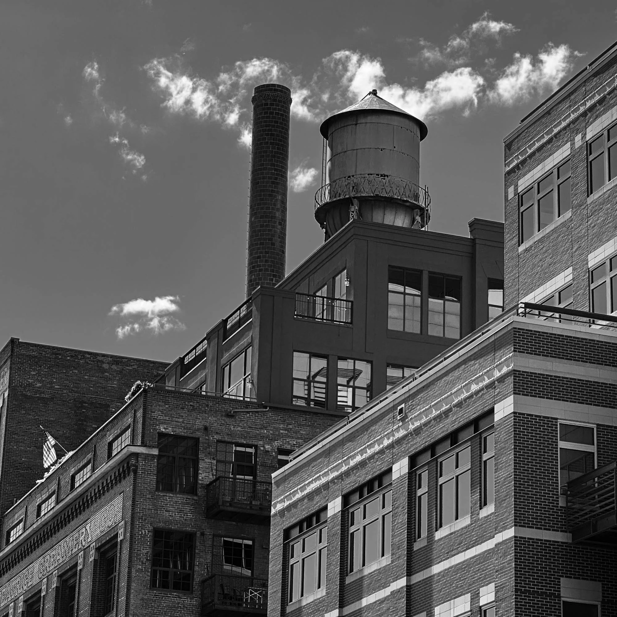 Black and white image of a cityscape showing multiple brick and glass buildings with water towers and a brick chimney on top. The sky is partly cloudy.