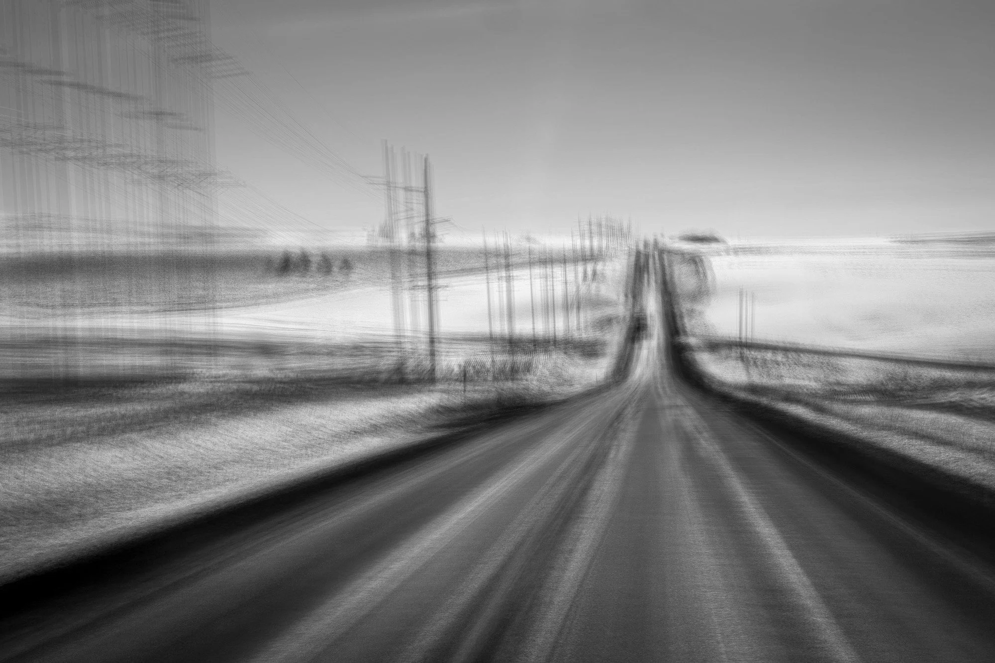 A black and white photo of a road leading through a rural area with power lines on both sides, appearing blurred and elongated to create a motion effect.
