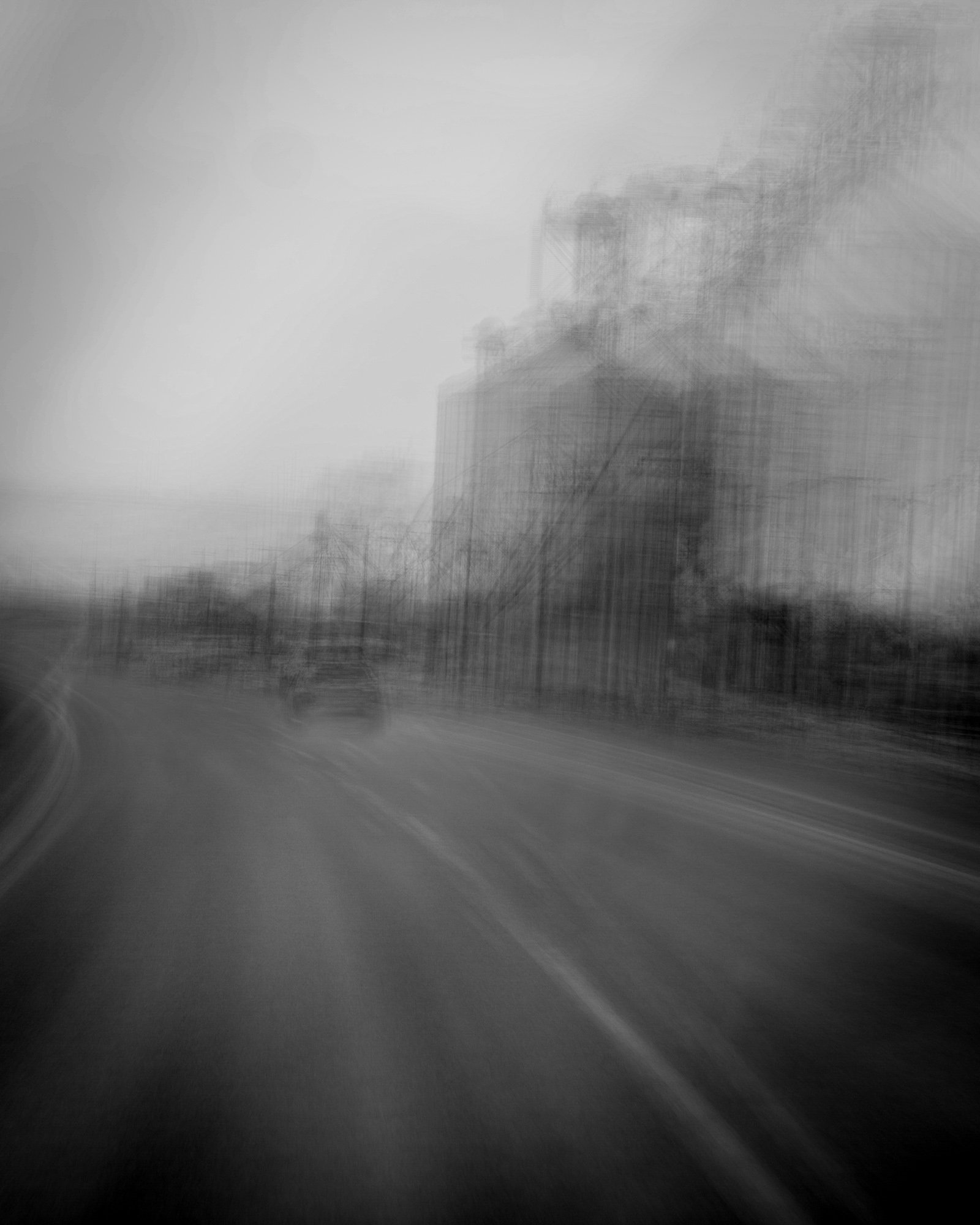 Blurred black and white photo of a road with buildings in the background.