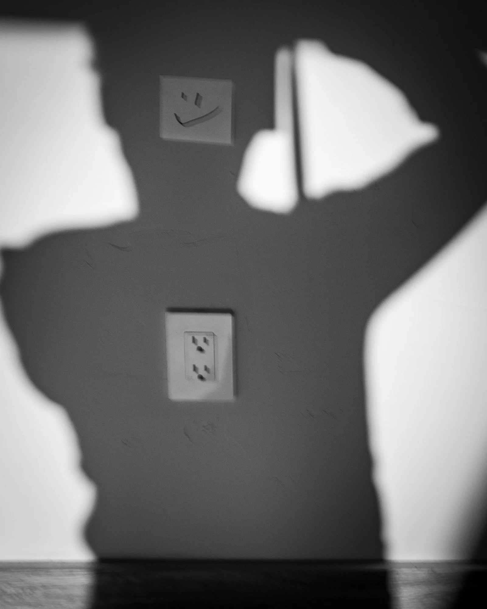 Shadow of a plant on a wall with an electrical outlet and a paper with a smiley face drawing in the background.