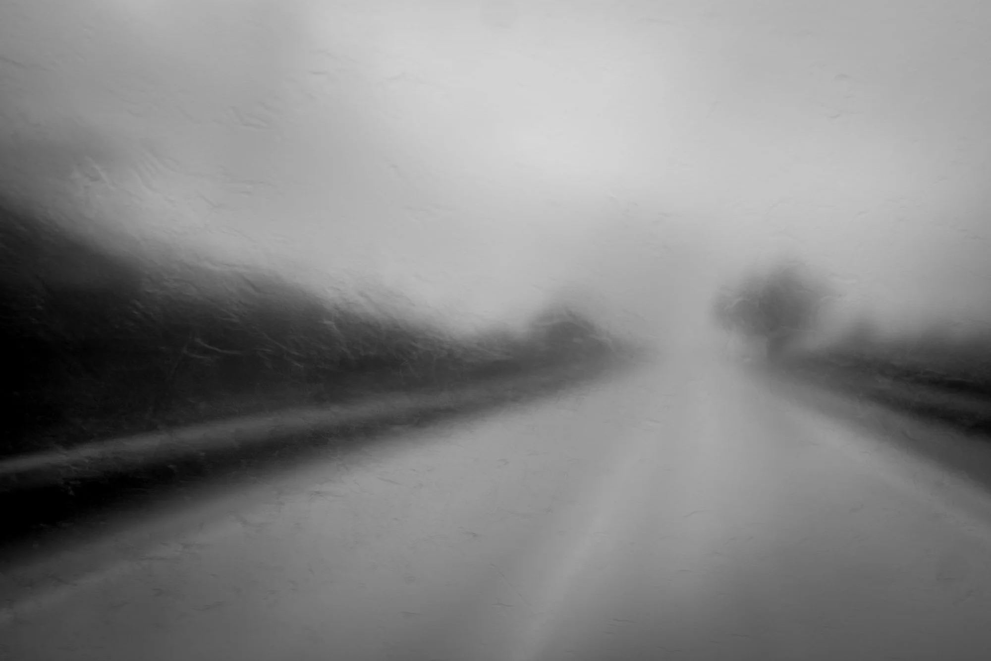 A black and white blurred photograph of a road or highway with foggy or rainy weather, causing low visibility.