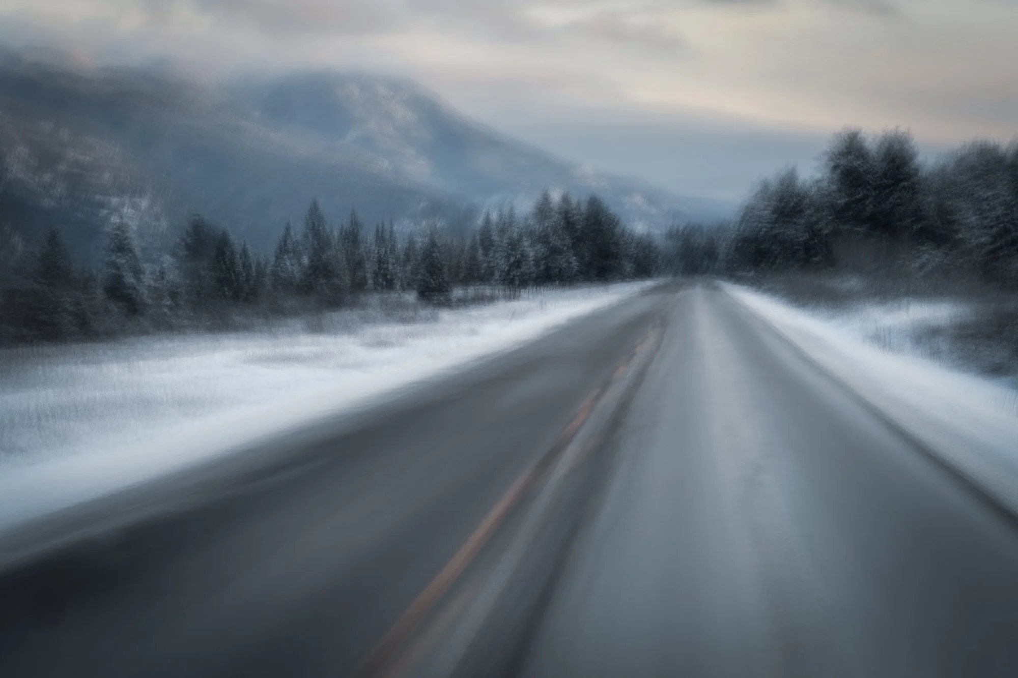 A blurred winter scene of a two-lane road with snow on the ground, flanked by trees and mountains in the distance, under cloudy skies.