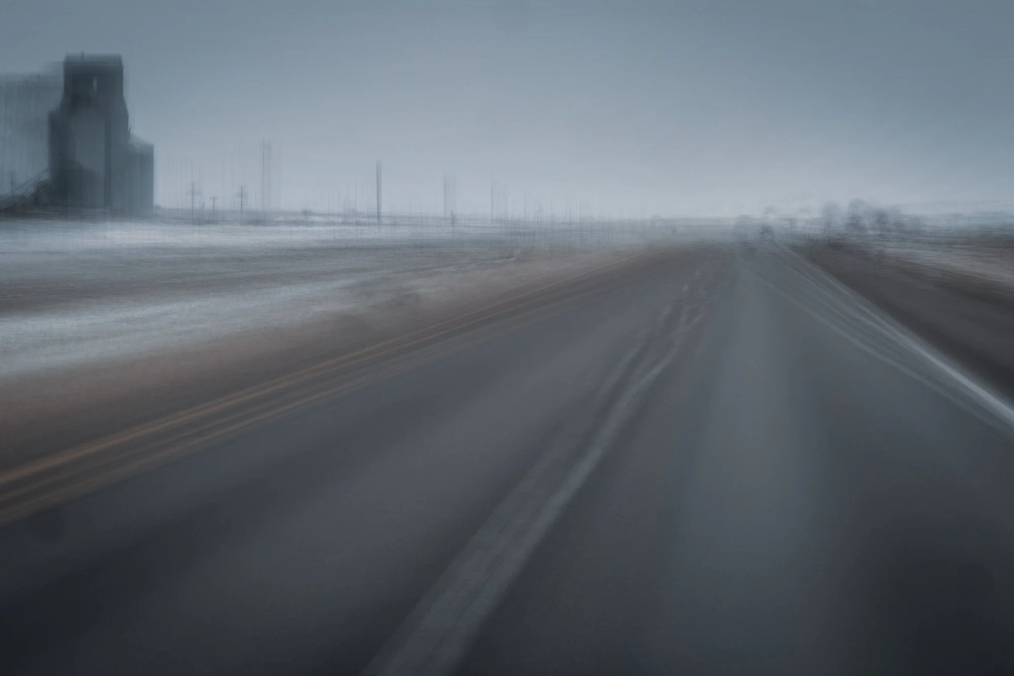 Blurred view of a wet road and distant buildings under a gray sky, taken from a moving vehicle on a highway.