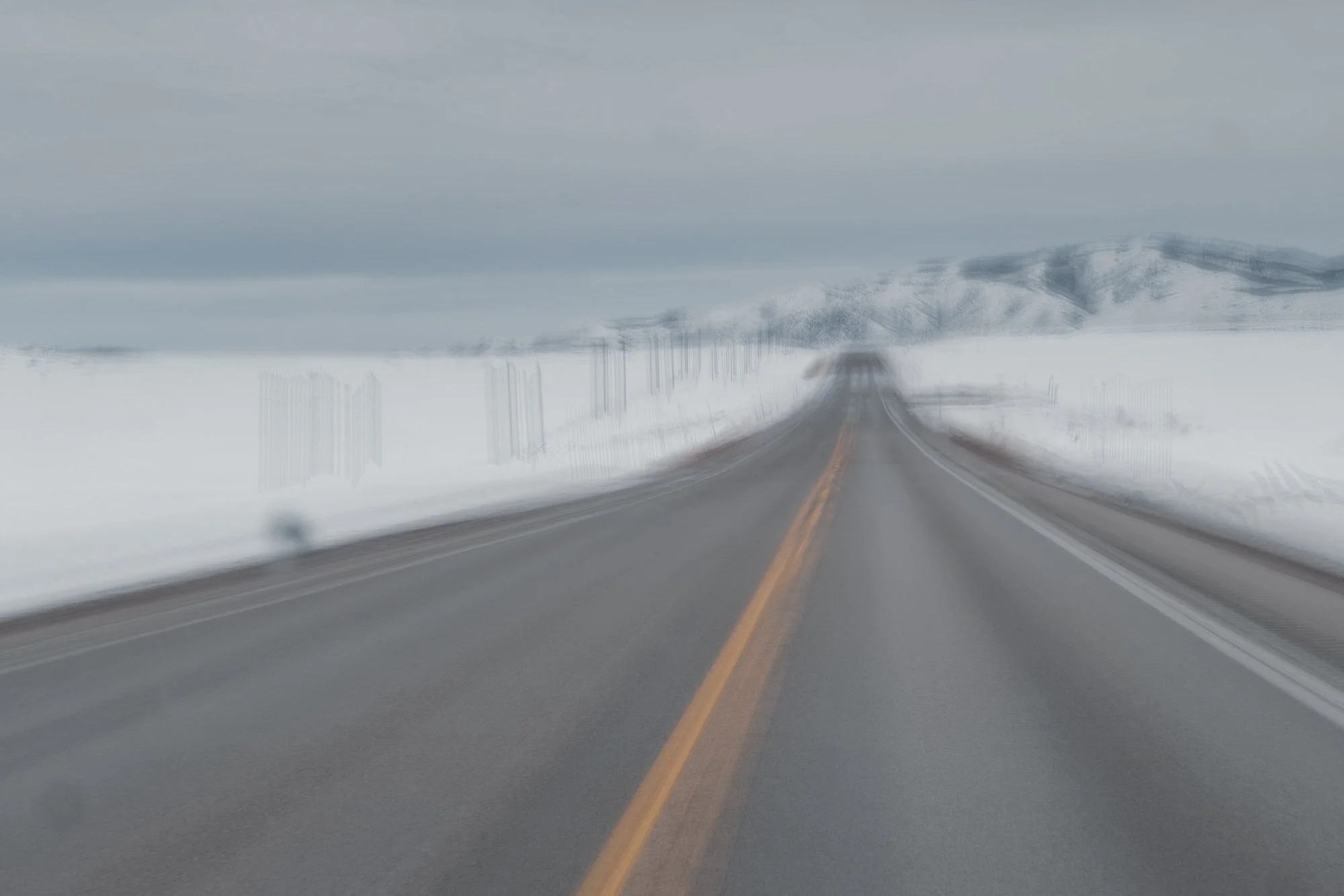 A highway stretching into the distance with snow-covered fields on either side and mountains in the background. The road has a yellow centerline and appears to be slightly blurred.