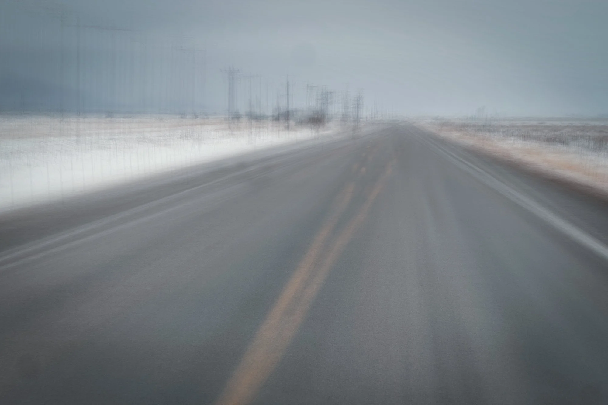 Blurred photo of a two-lane road with yellow dividing lines, extending into the distance through a flat, snowy landscape with power lines on the sides.