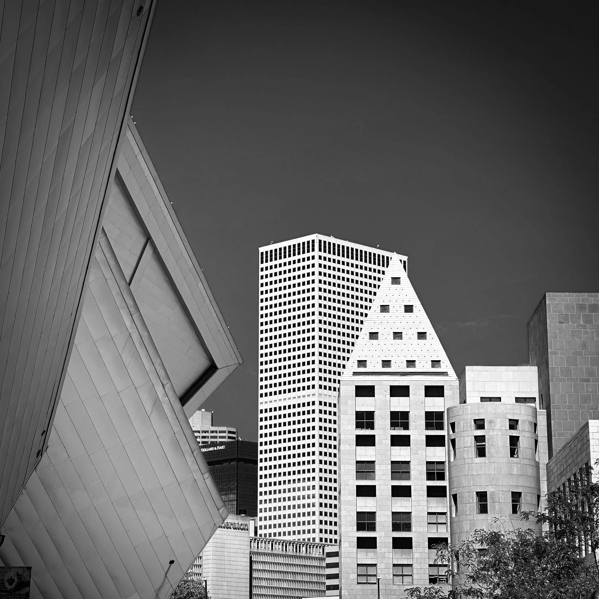 Black and white photo of various modern buildings in an urban cityscape, including a tall rectangular grid-patterned skyscraper, a pyramid-shaped building with small windows, and other geometric structures, with a clear sky in the background.