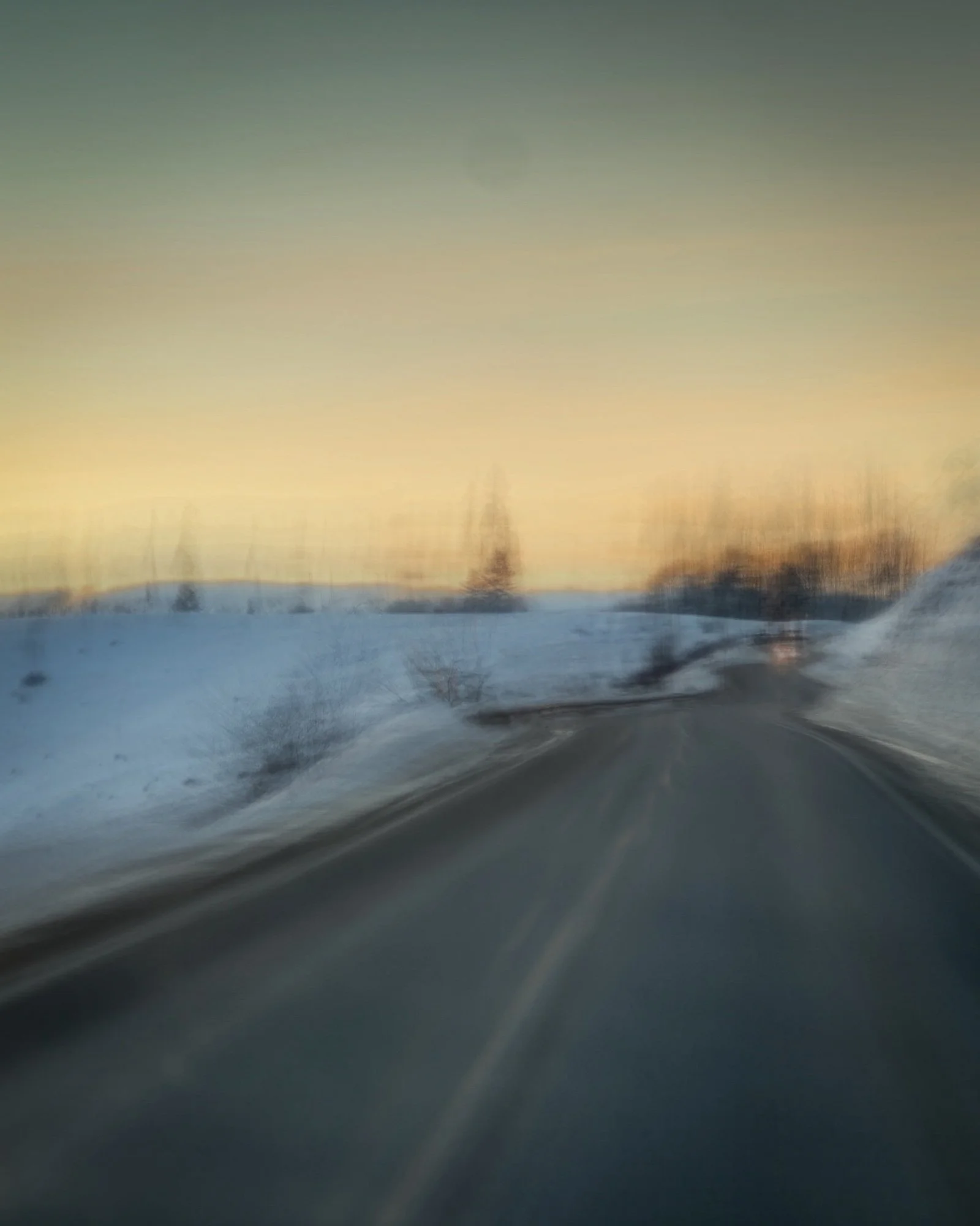 Blurred winter landscape viewed from a moving car, showing snow-covered ground, trees, and a sunset or sunrise sky.