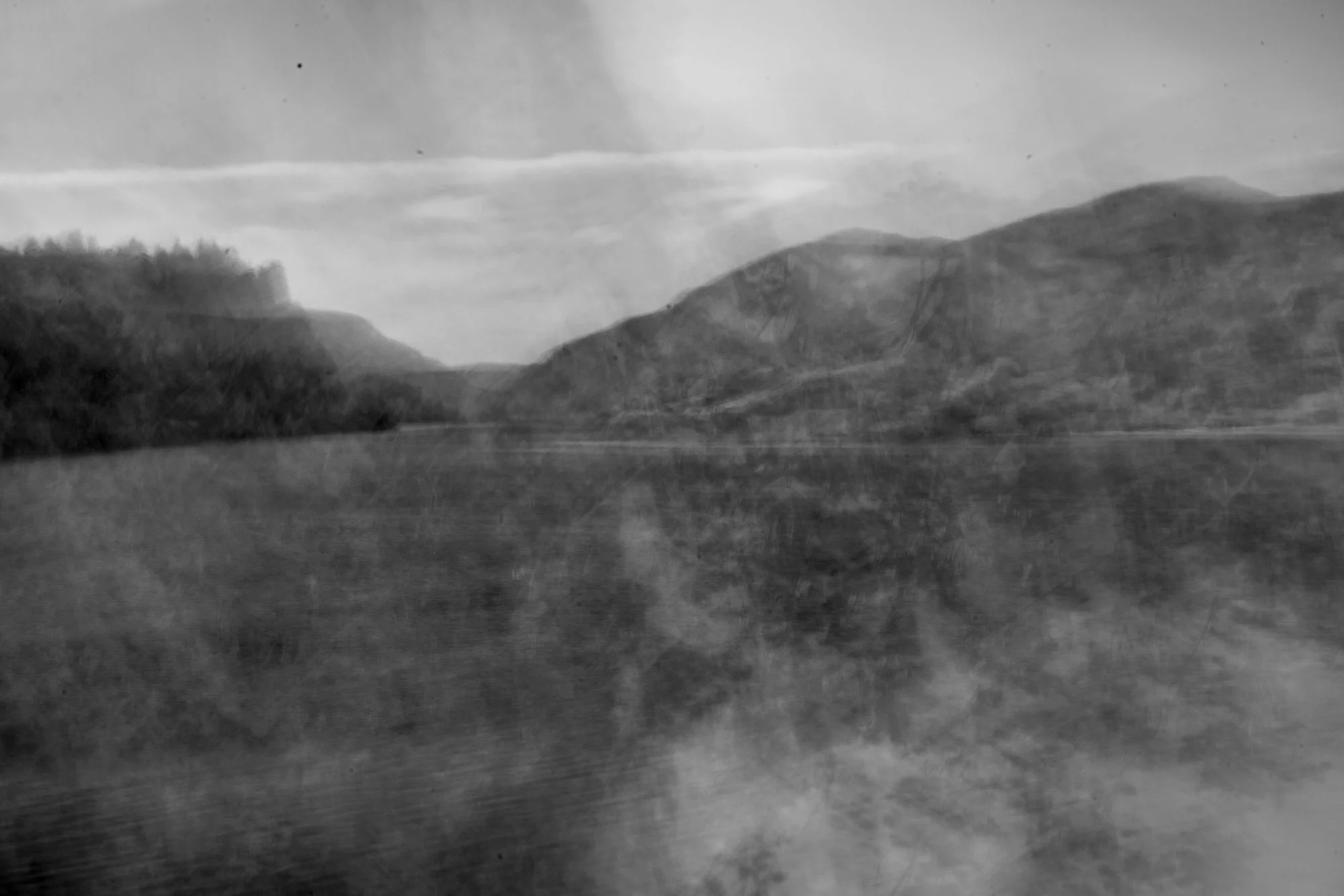 A black-and-white photograph of a river surrounded by mountains with a cloudy sky.