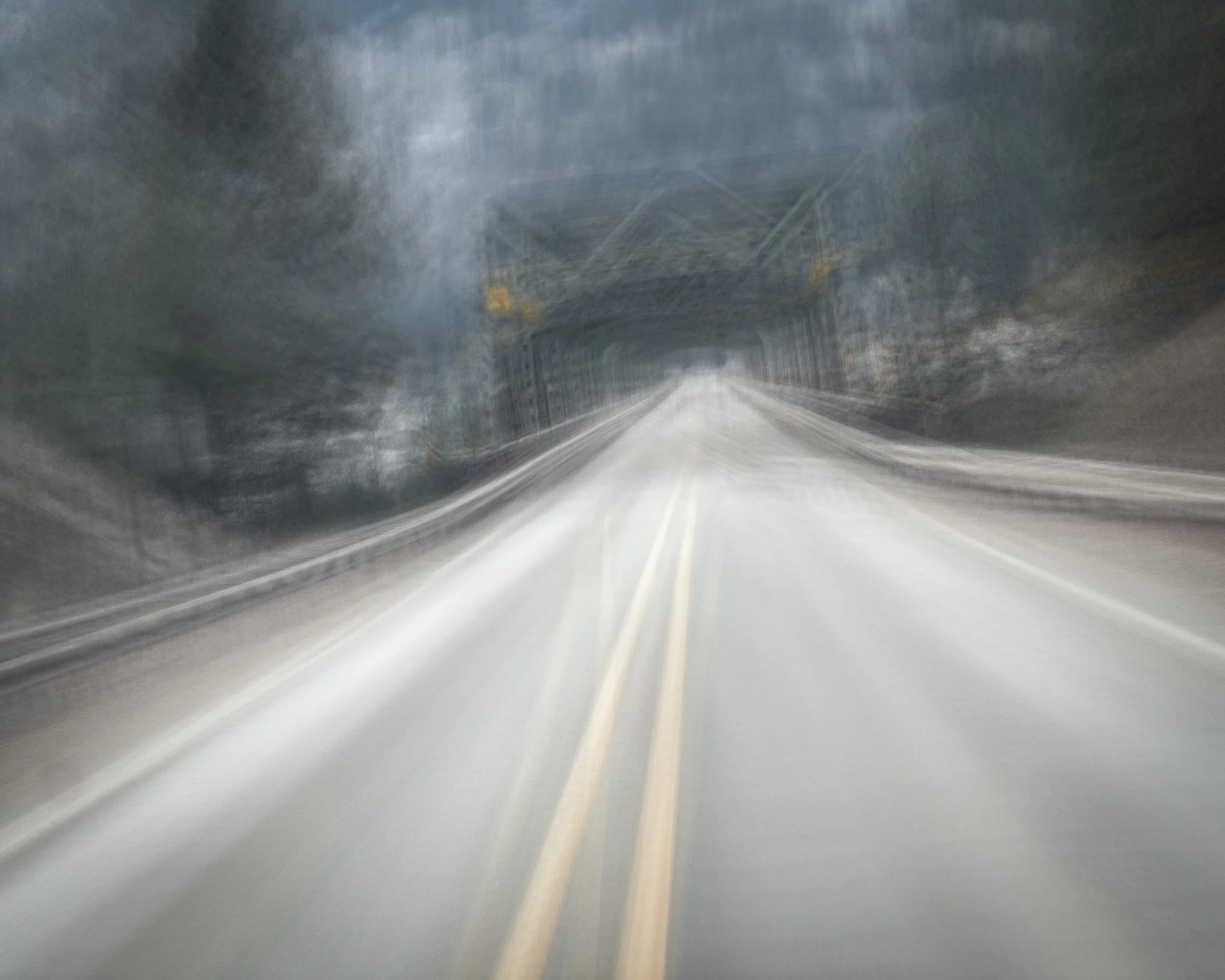 A blurry image of a road leading to a bridge, with trees on both sides and a dark sky overhead.