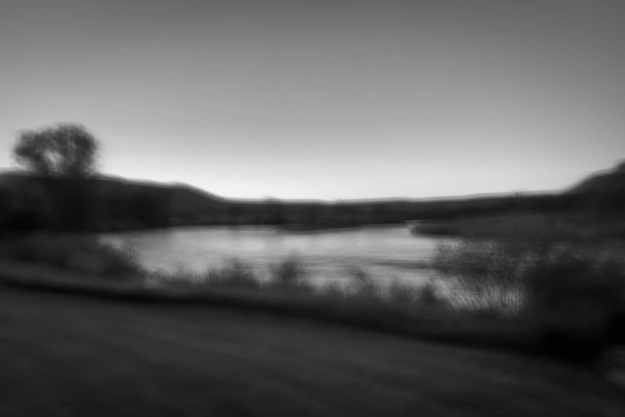 Blurry black and white landscape photo of a river flowing through hills with trees, captured at dusk or dawn.