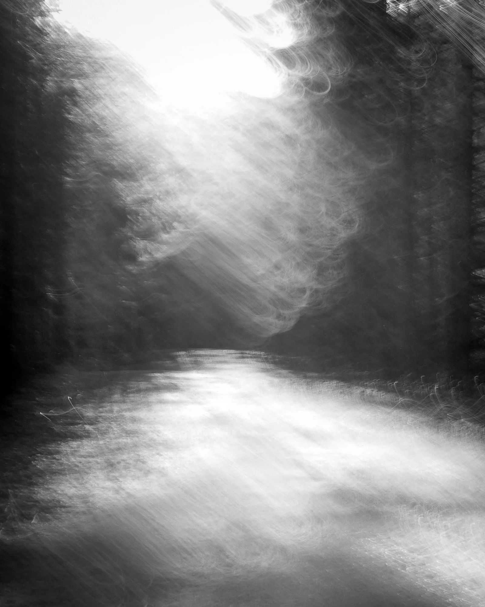 Black and white photograph of a river with blurred trees and sky reflecting on the water, with bright sunlight creating lens flare.