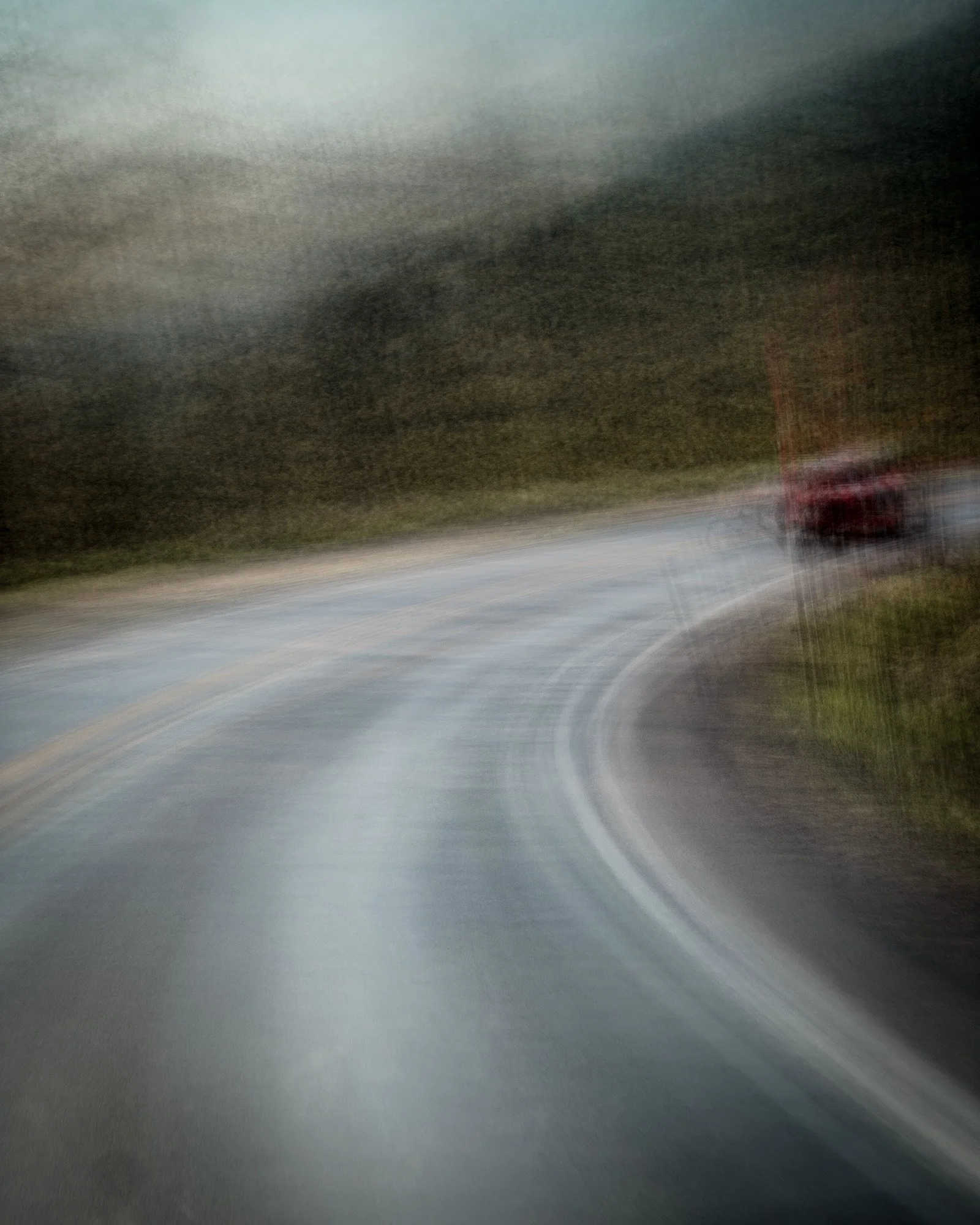Blurry photo of a winding road with a red vehicle on the right side, surrounded by trees and a dark, cloudy sky.