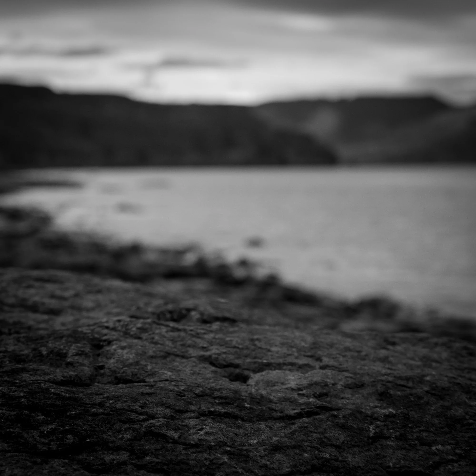 Black and white photo of a rocky shoreline with water and distant hills in the background.