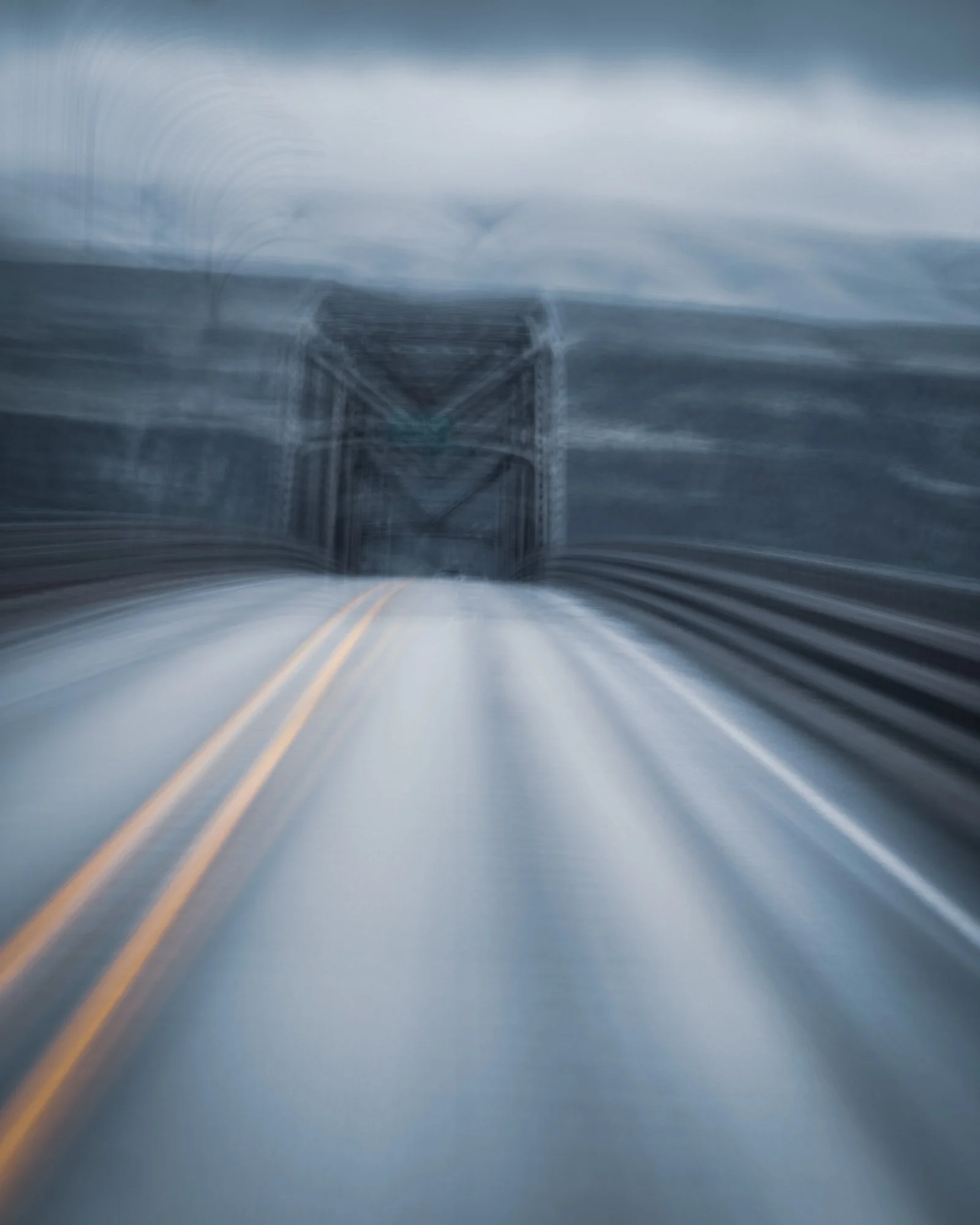 Blurred view of a bridge over a body of water with skies overhead, as seen from a moving vehicle.