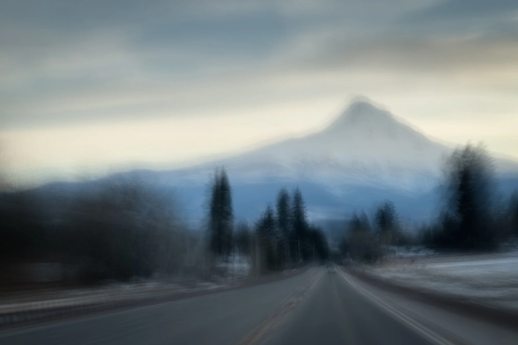 Blurry road leading towards mountains with a prominent peak and trees on either side, under a cloudy sky.